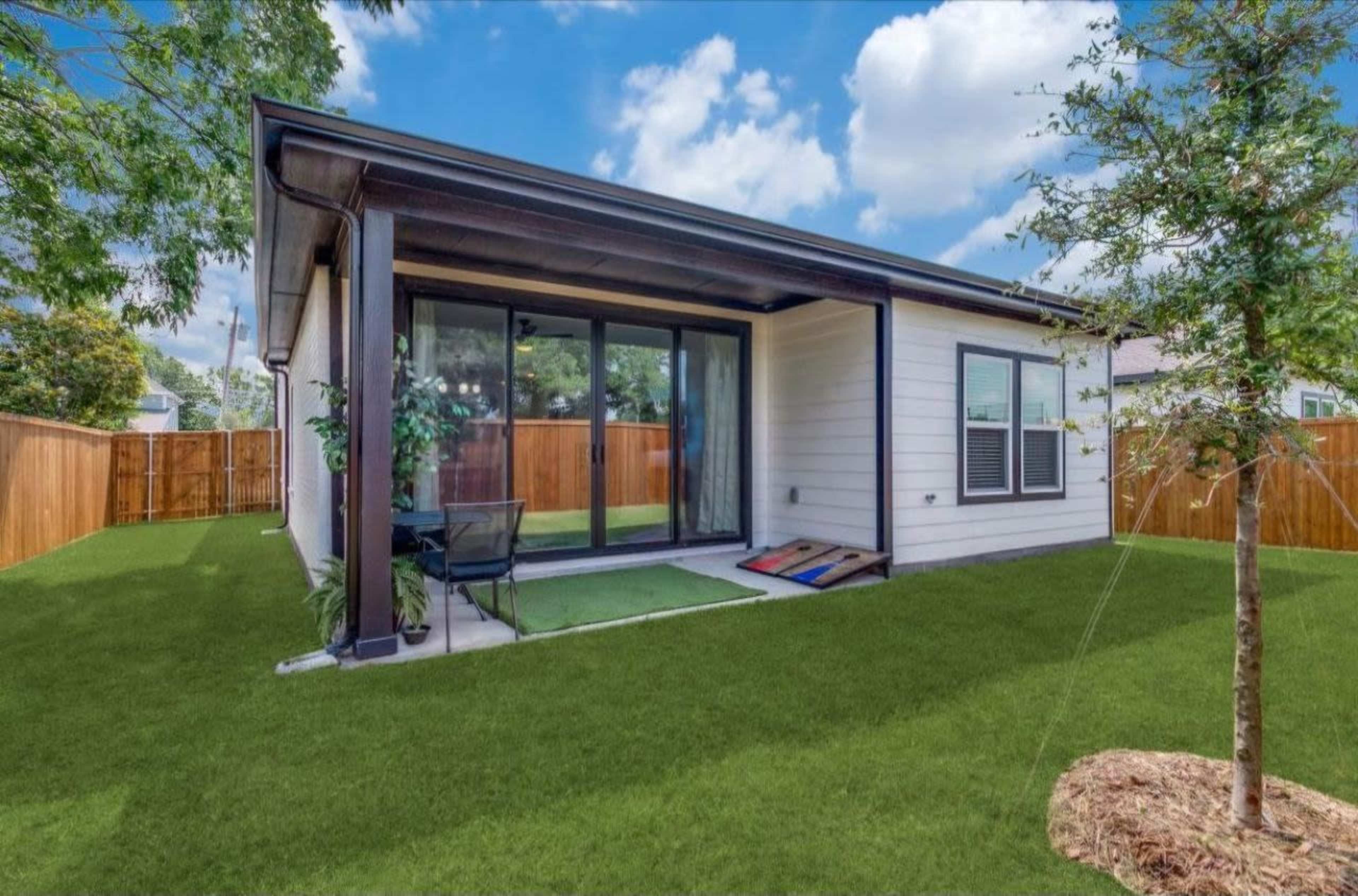 A small modern house features a patio area with sliding glass doors opening to a grassy backyard surrounded by a wooden fence.