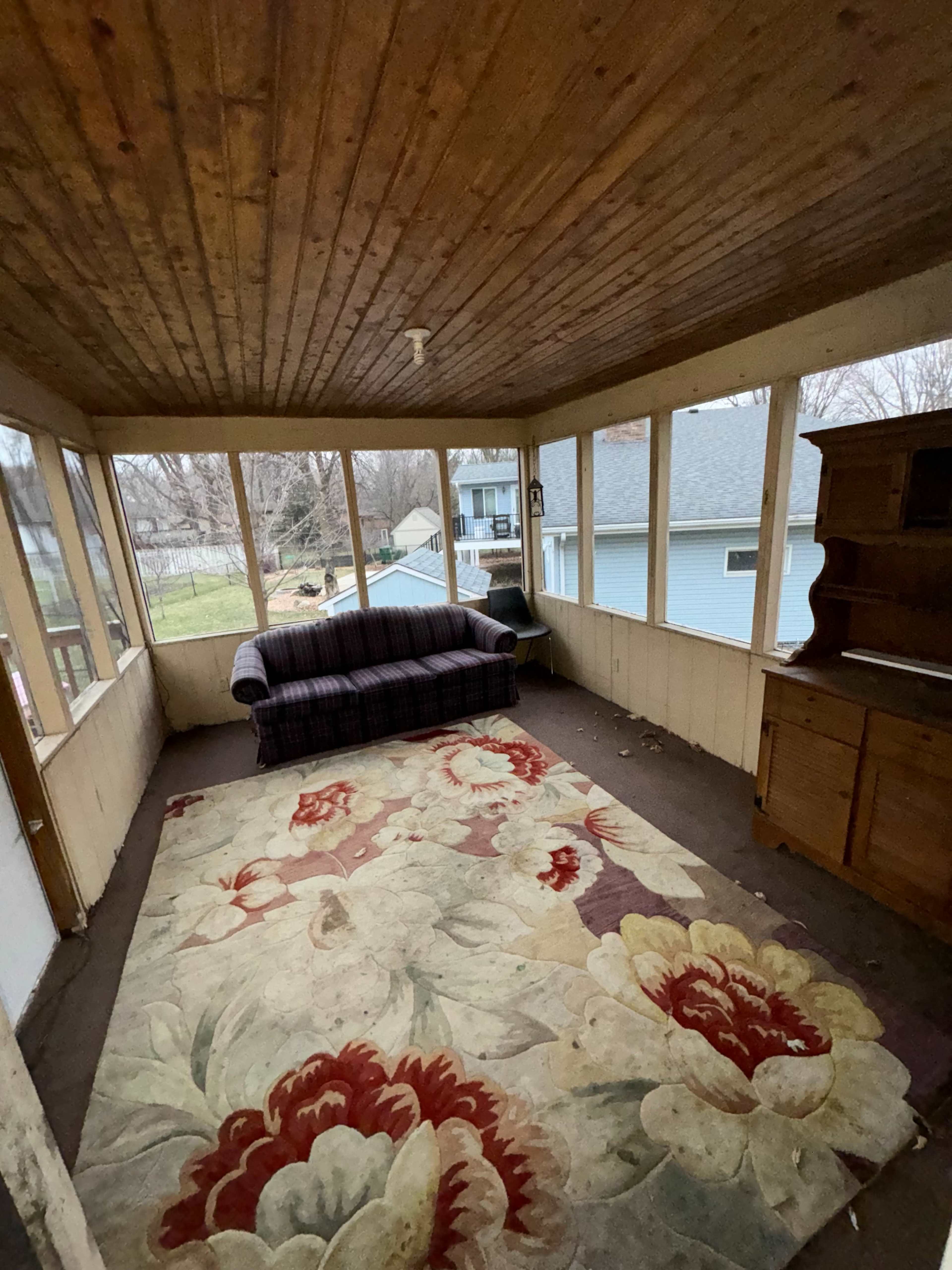 A screened-in porch featuring a plaid sofa, a floral rug, and a wooden cabinet, with windows providing a view of the outside.