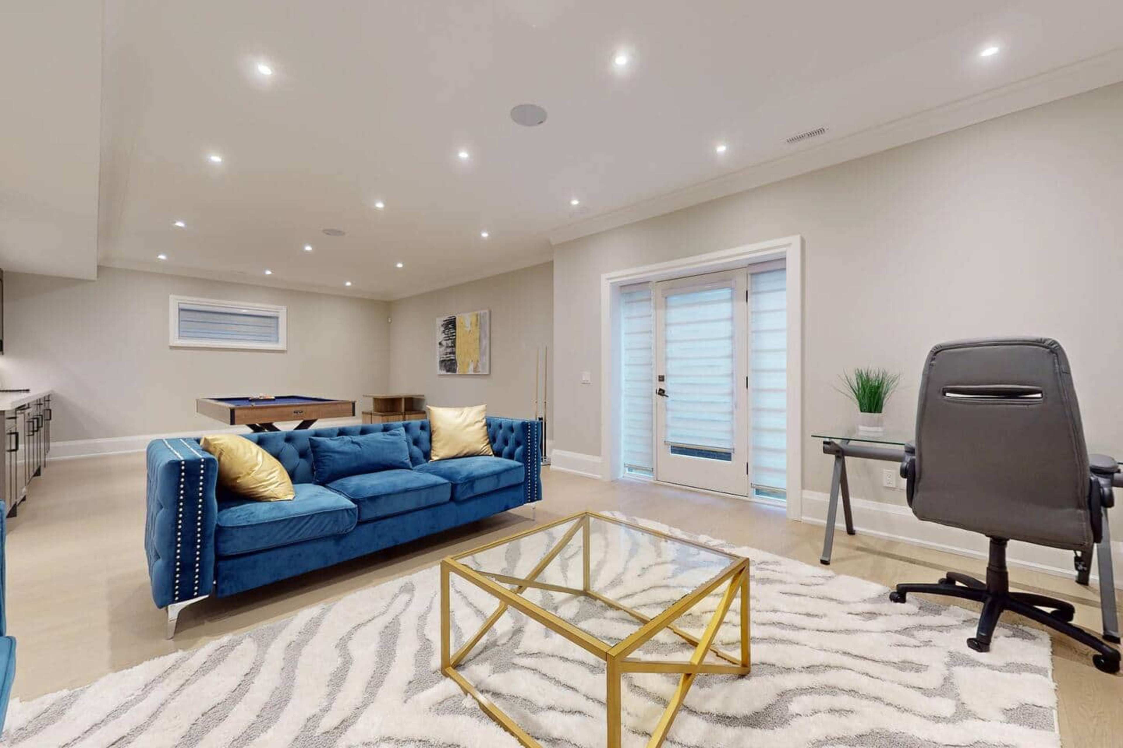 The image features a modern living space with a blue sofa, a glass coffee table, and an office chair next to a desk, all illuminated by recessed lighting.