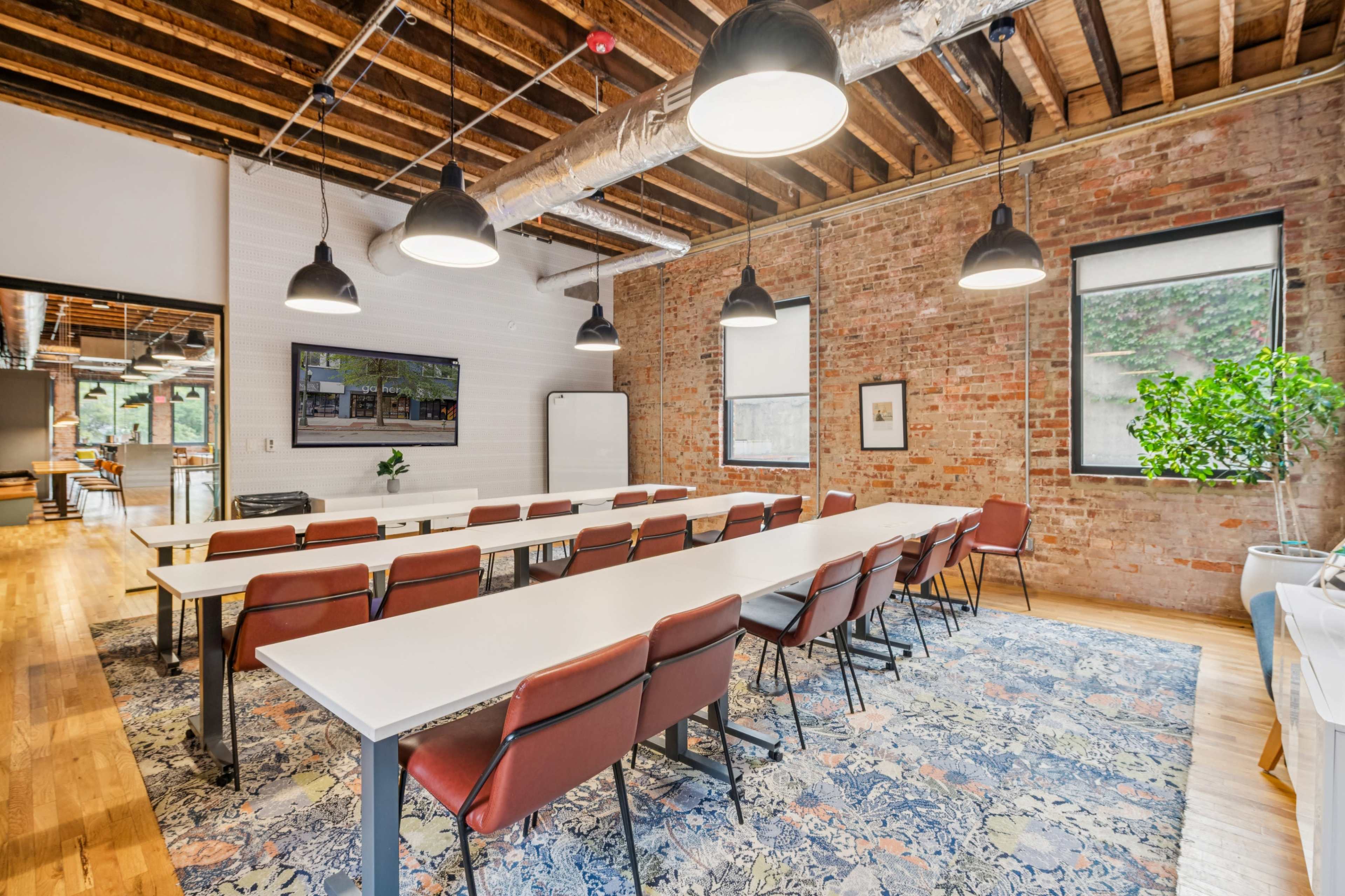 The image shows a modern conference room with rows of white tables and red chairs, set against a backdrop of exposed brick walls and wooden beams, with overhead lighting and a large screen on the wall.