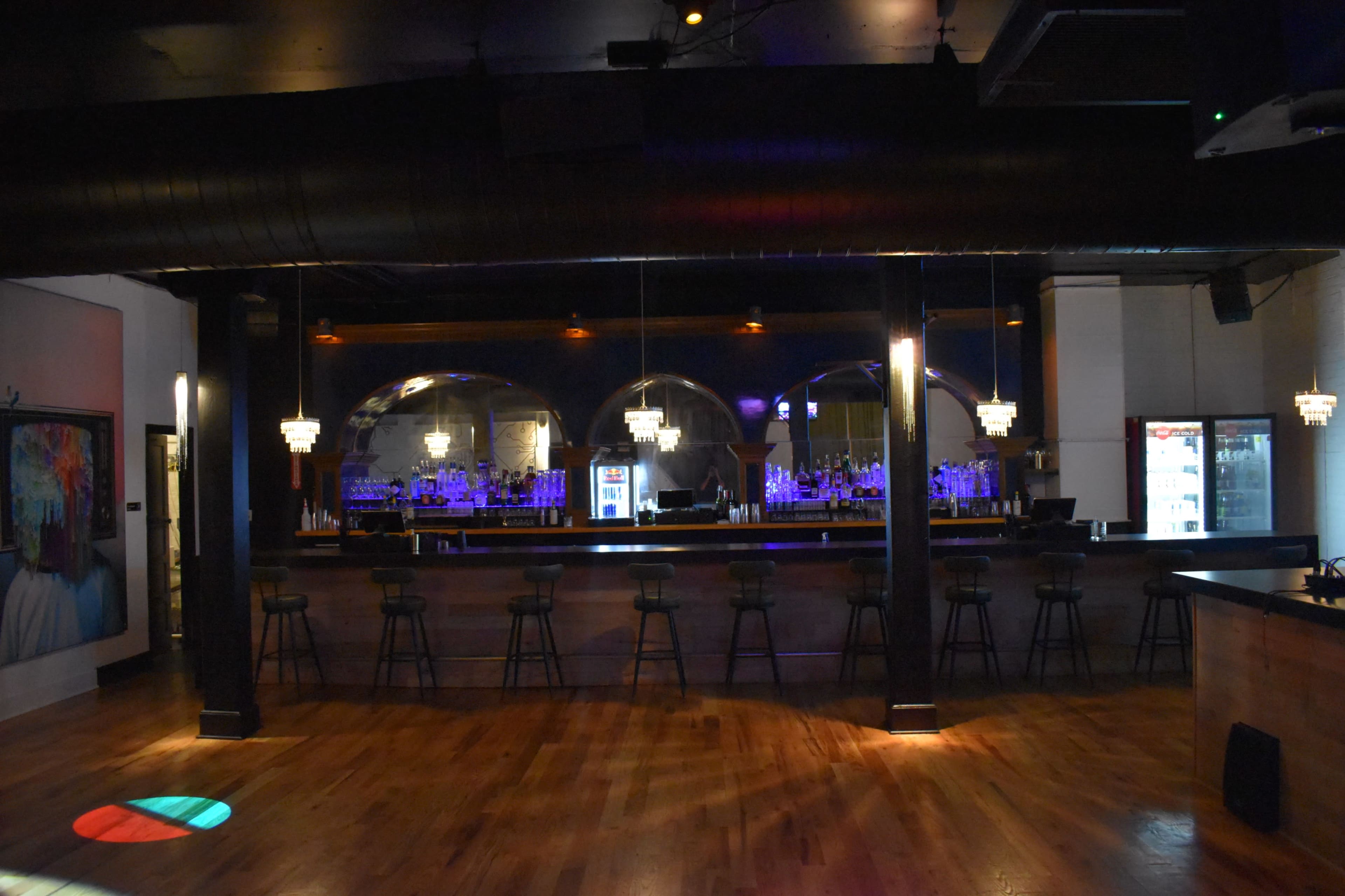 A dimly lit bar area with a long wooden counter, high stools, and a backlit shelf displaying various bottles behind the bar.