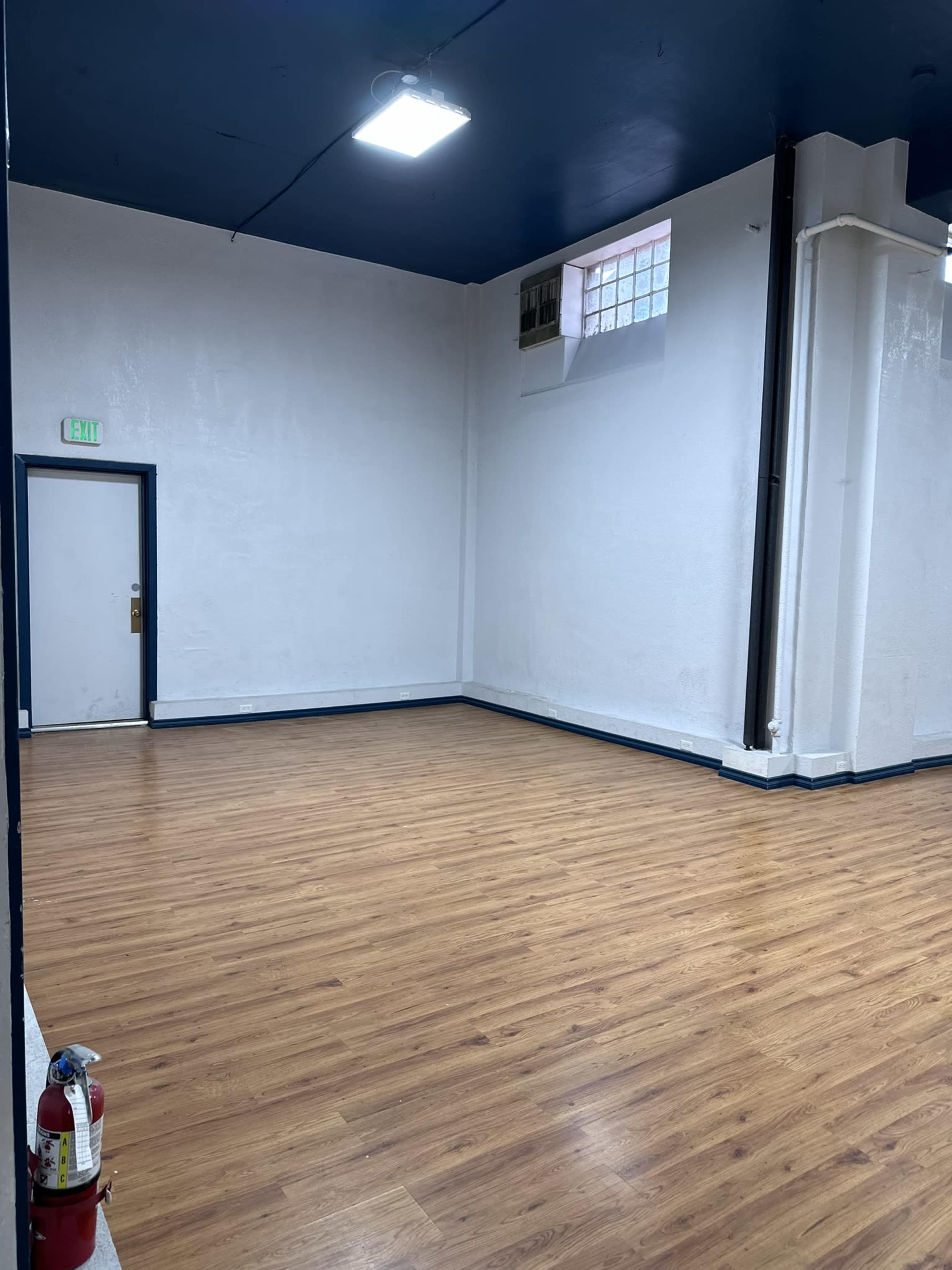 A spacious, empty room with wooden flooring and white walls, featuring a door on one side and a fire extinguisher mounted near the entrance.