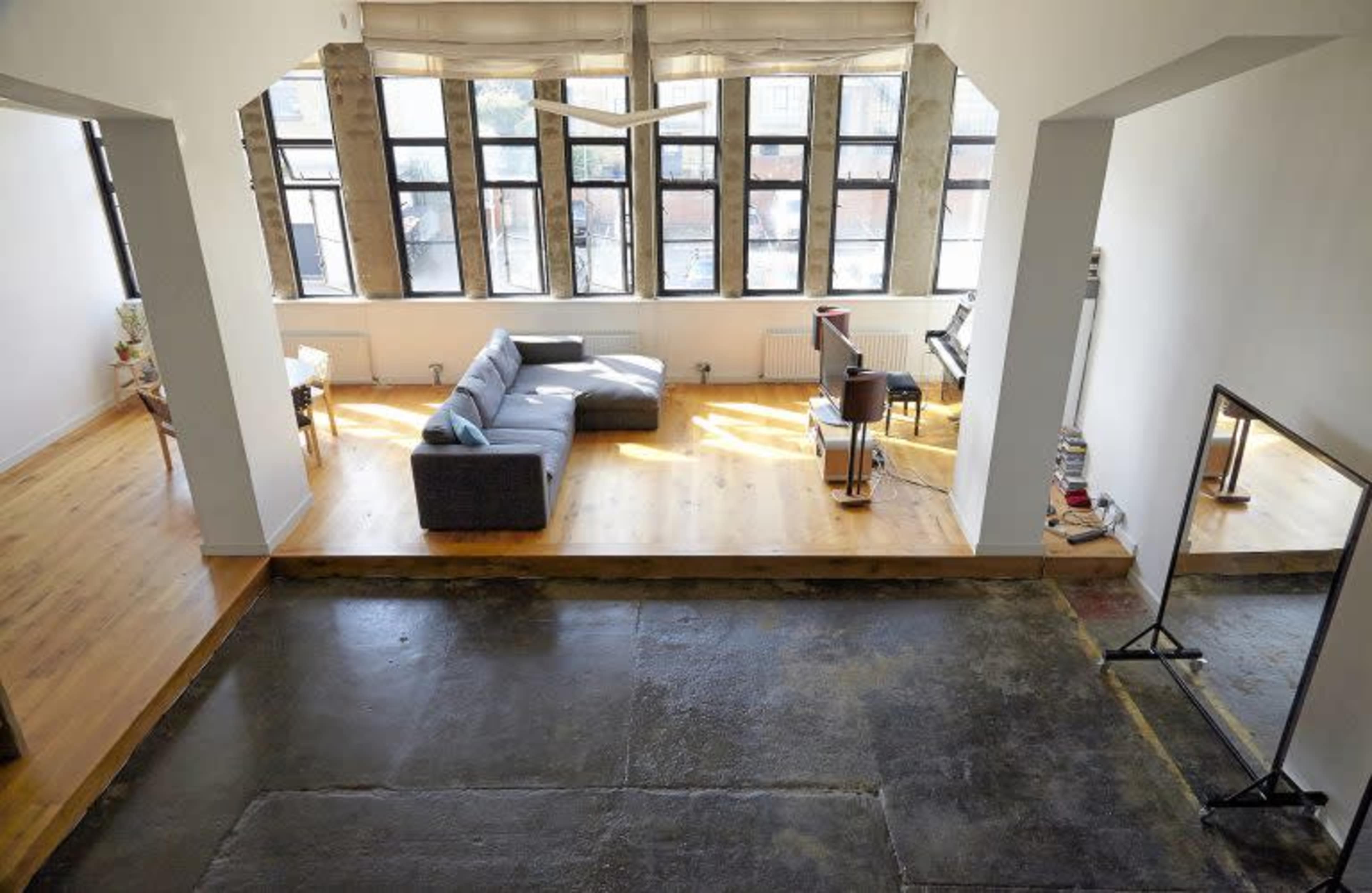 The image shows a spacious loft-style room with large windows, featuring a gray couch, a coffee table, and a small music setup in the corner.