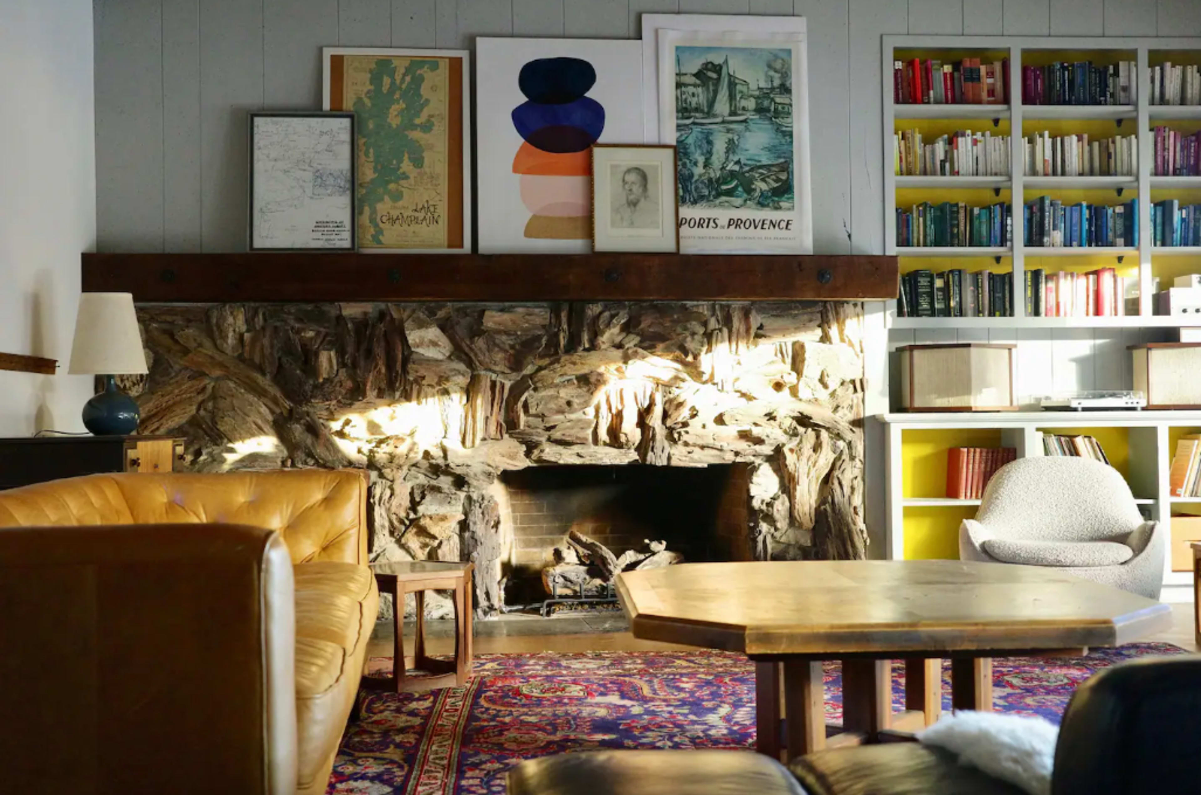 The scene shows a cozy living room with a stone fireplace, a wooden coffee table, and framed artwork on the walls, surrounded by bookshelves filled with books.
