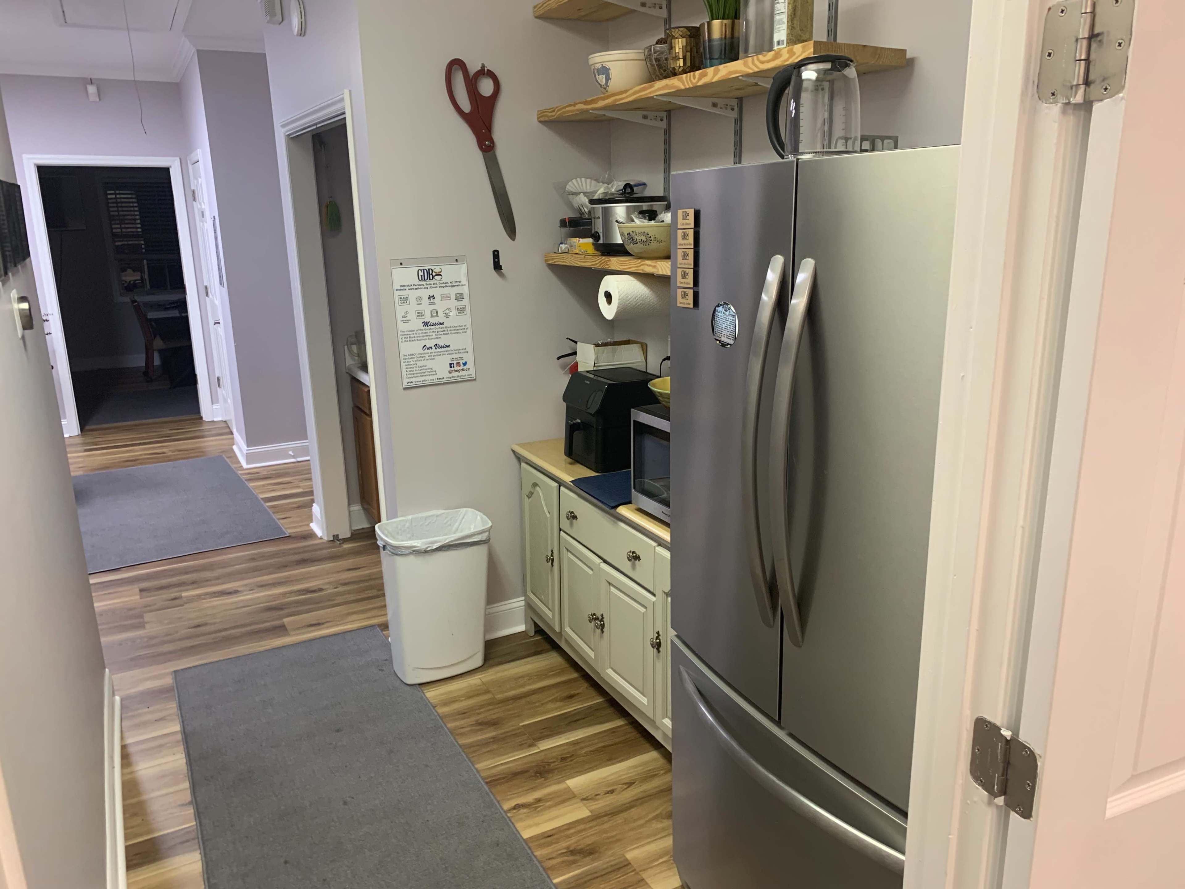 The image shows a hallway leading to a kitchen area with a stainless steel refrigerator, a countertop with utensils, and a trash can on the floor.