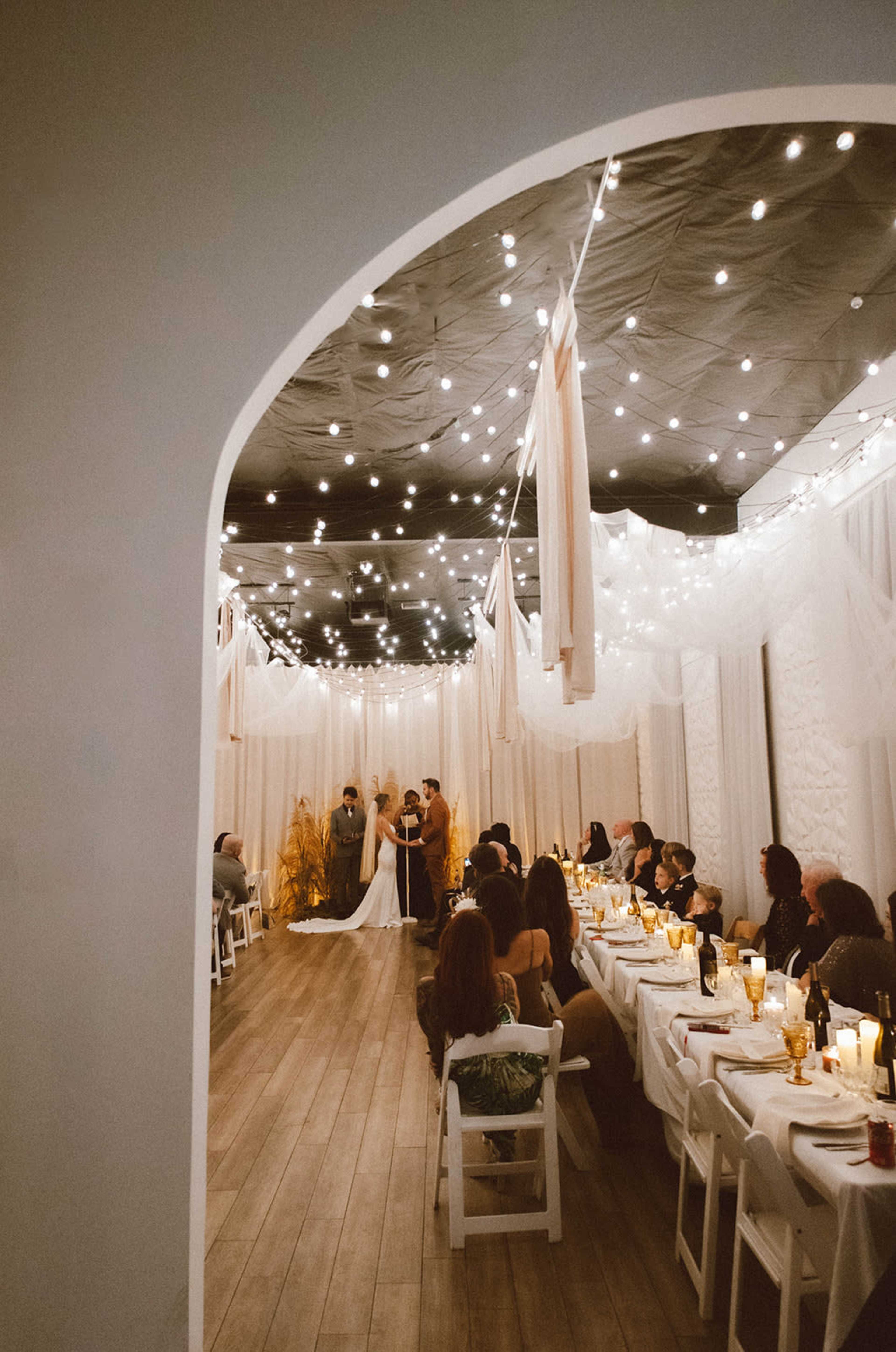 A wedding ceremony takes place under string lights in a decorated indoor space with guests seated at long tables.