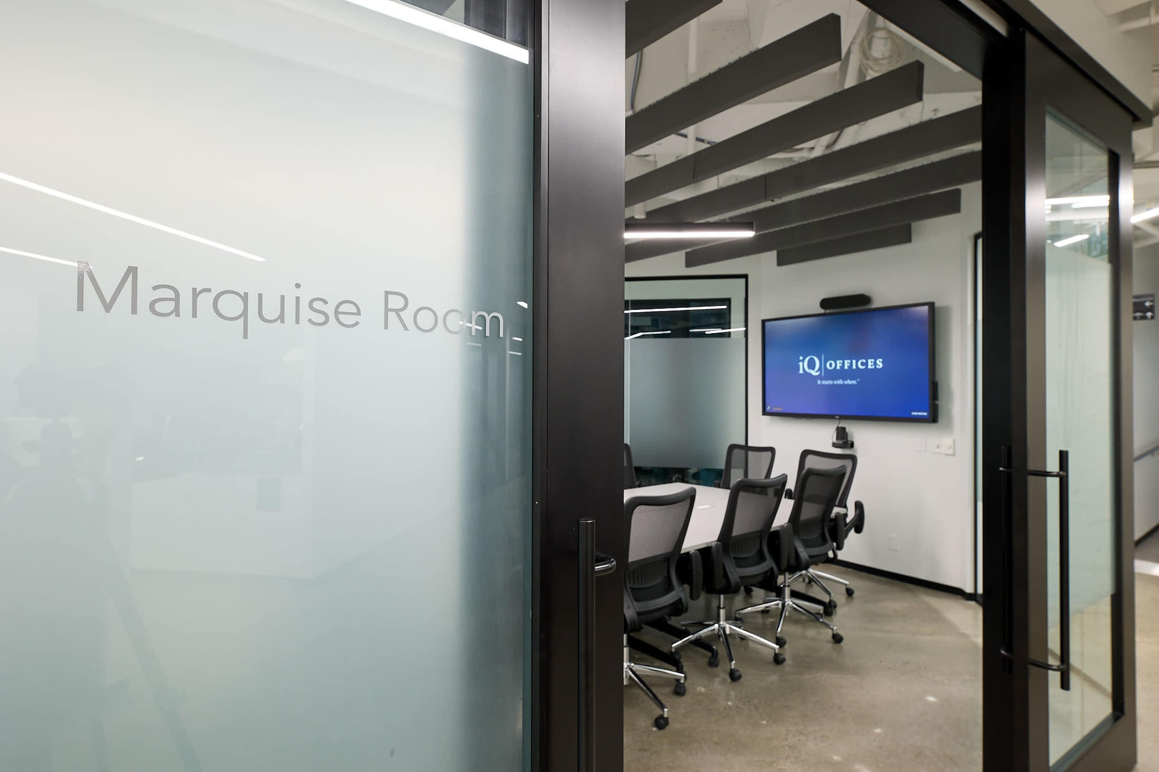 The image shows a modern conference room with glass walls, featuring a large screen displaying the "iQ Offices" logo and a nameplate indicating "Marquise Room."
