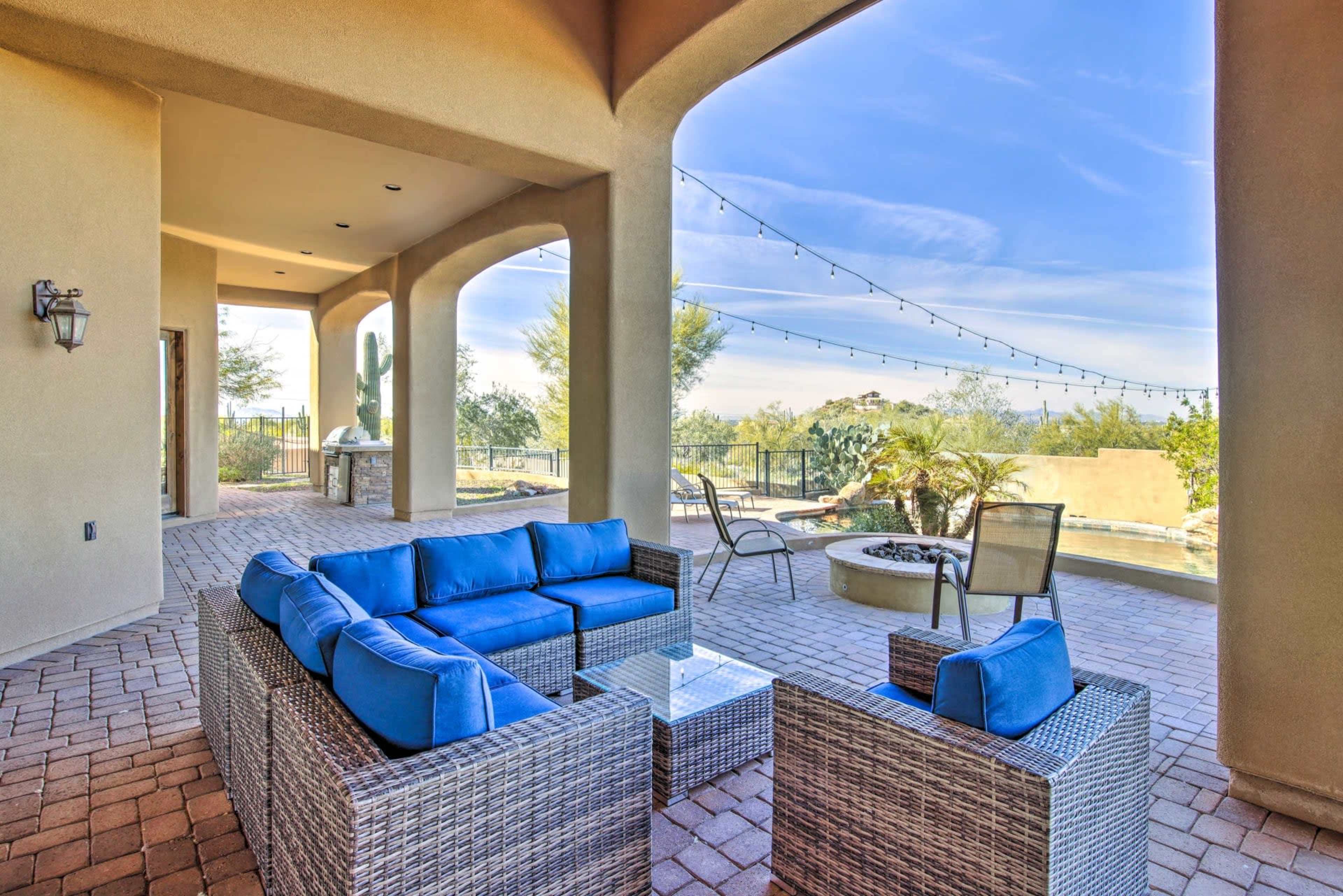 A patio area features woven furniture with blue cushions, a coffee table, and a view of a landscaped outdoor space with string lights overhead.