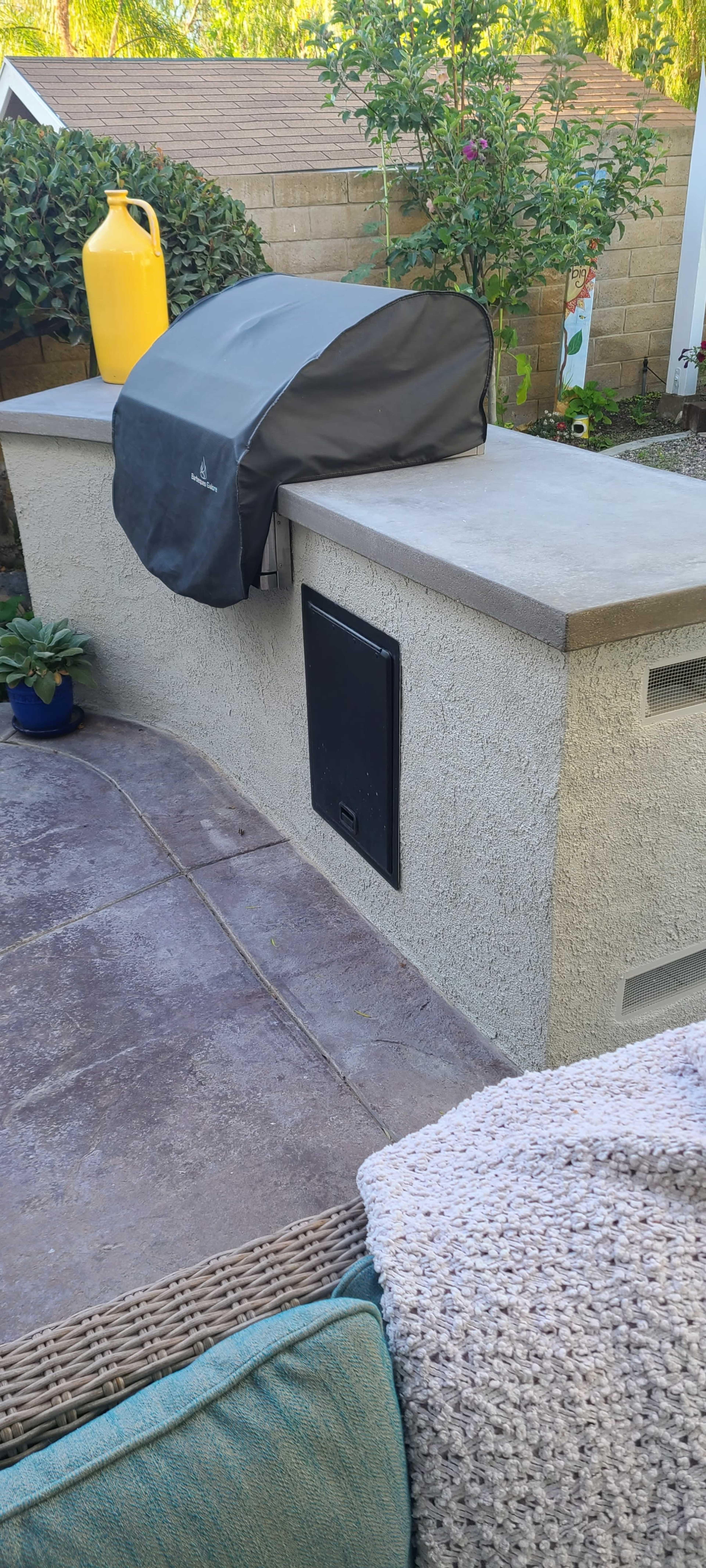 A grill is covered with a black cover on a concrete outdoor countertop next to a yellow container and plants.