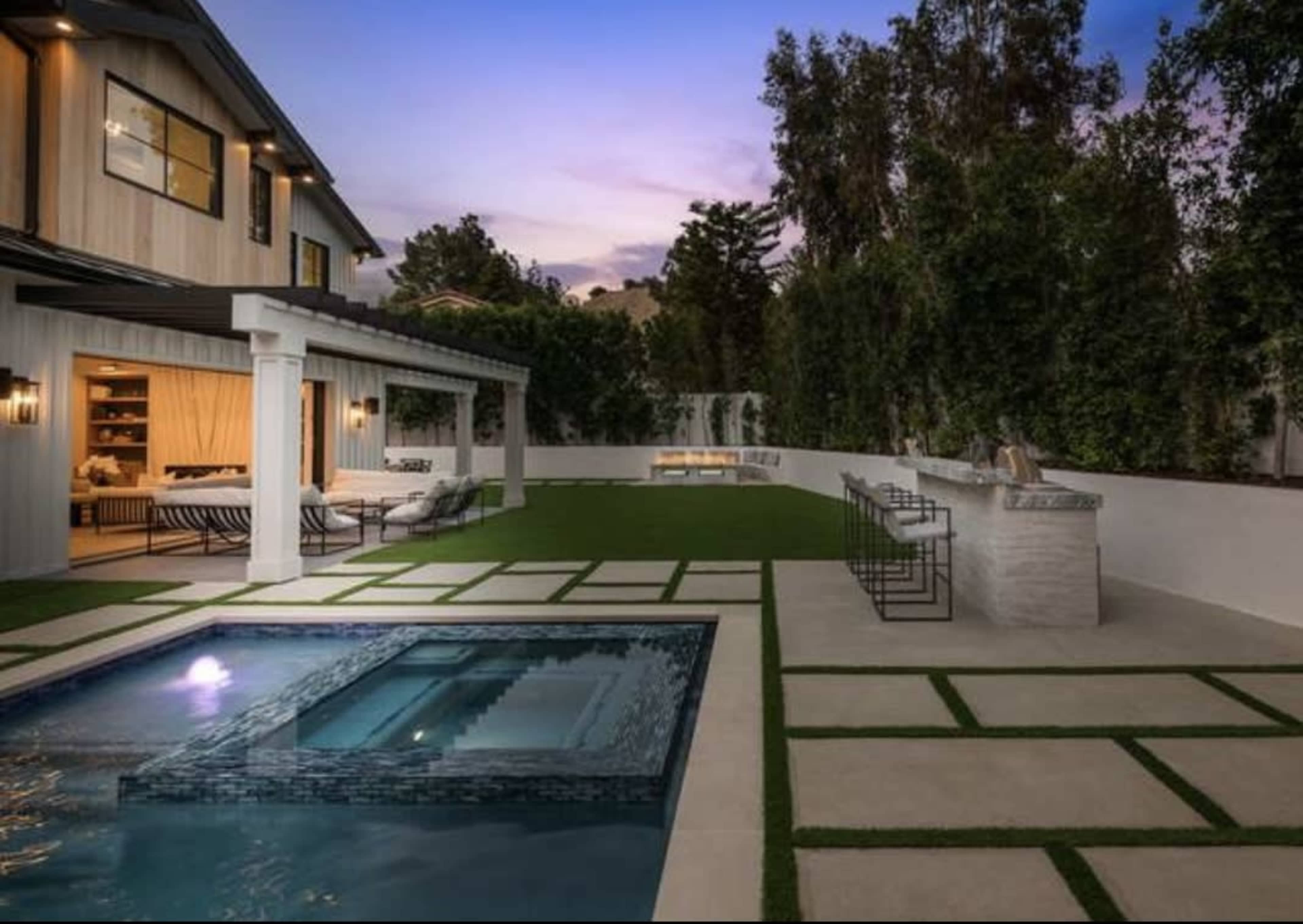 The image shows a modern backyard with a pool, patio seating area, and an outdoor bar set against a backdrop of trees and a house in twilight.