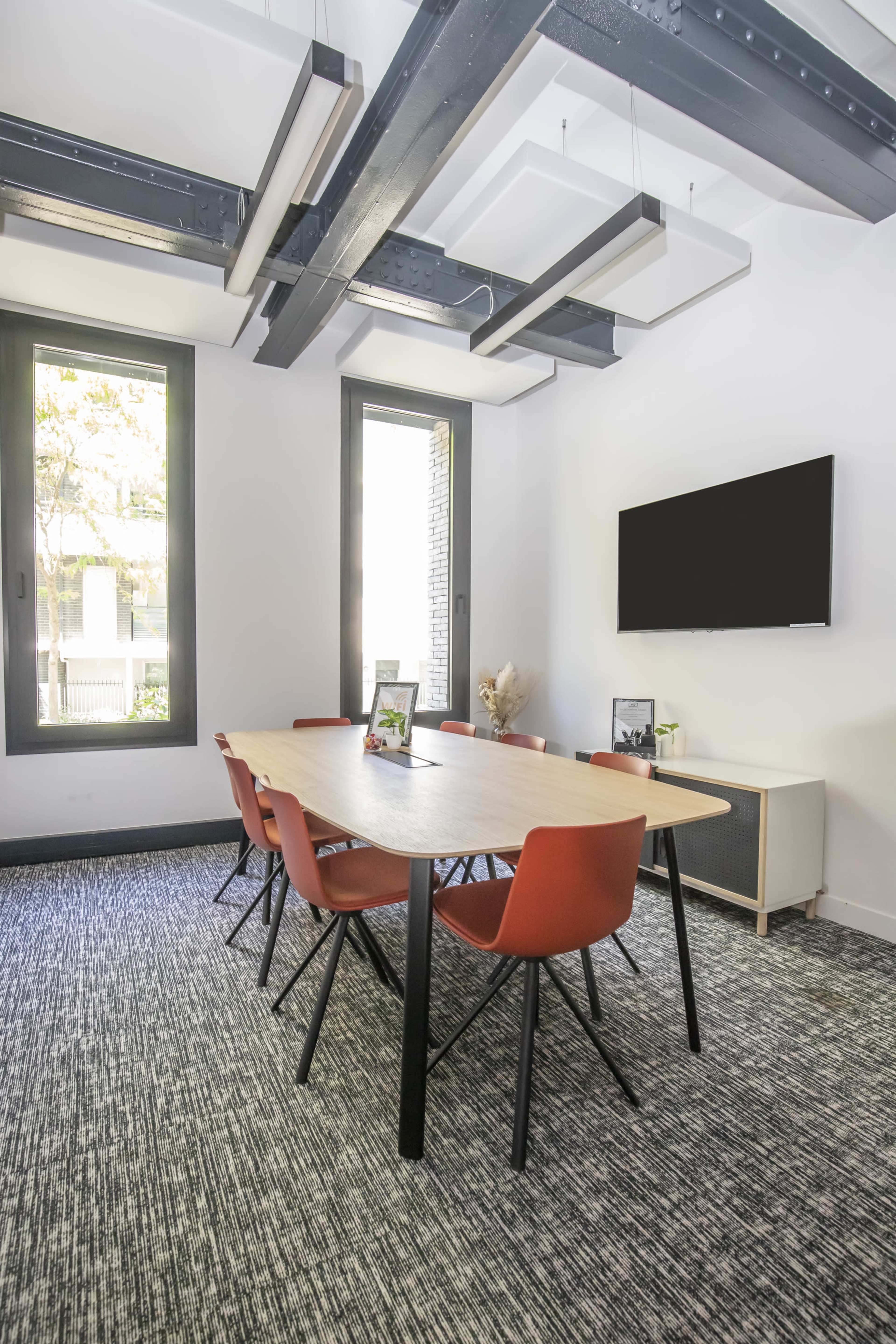 A modern meeting room features a large table surrounded by orange chairs, two windows, and a television mounted on the wall.