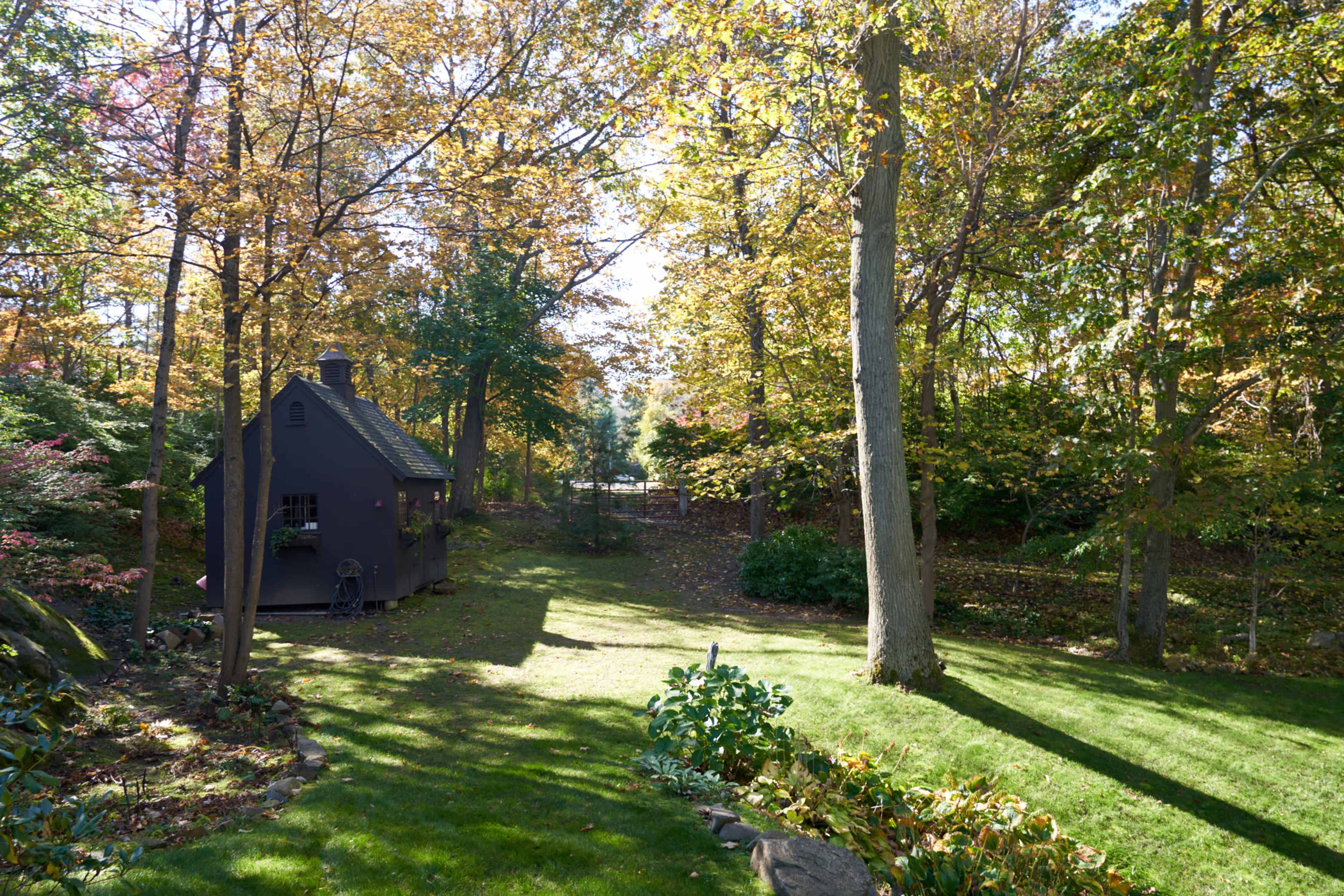 A small, dark-colored shed is situated in a sunlit garden surrounded by trees with autumn foliage.