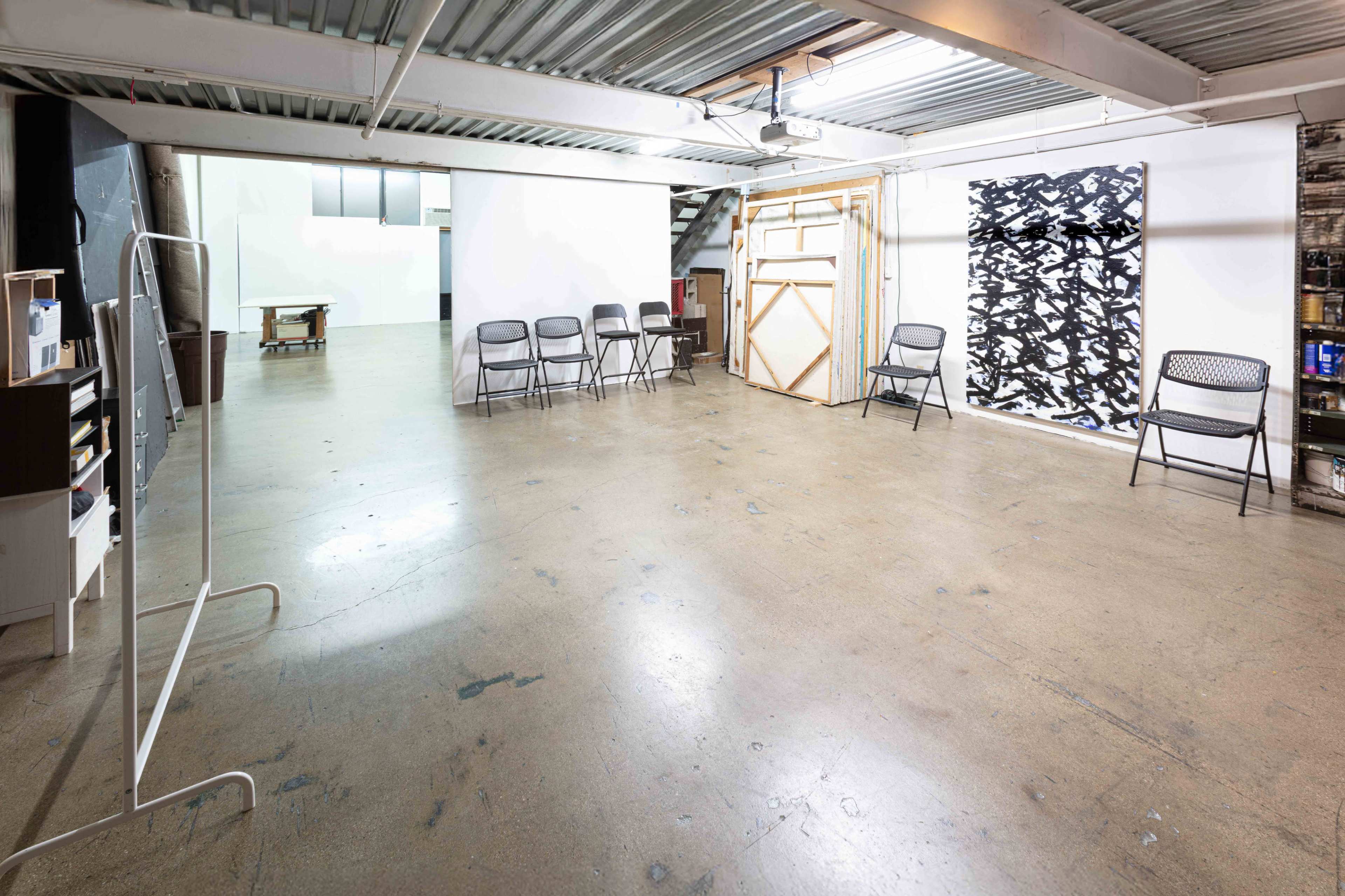 The image shows a spacious, unfinished interior with gray concrete flooring, several folding chairs arranged in a row, and a large abstract artwork on a wall.