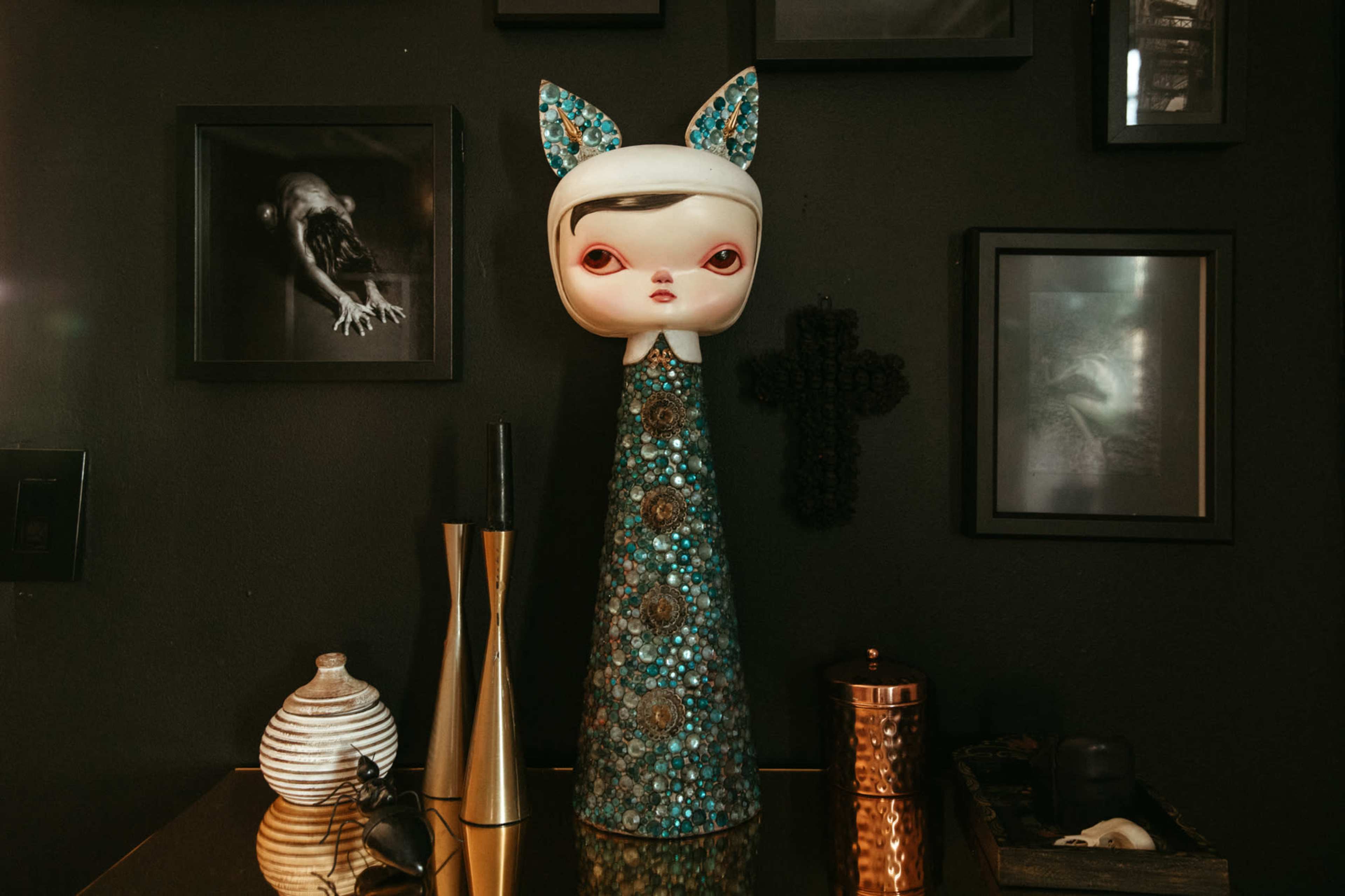 A tall, decorative doll with a patterned dress and cat-like ears stands on a shelf surrounded by various framed artworks and objects.