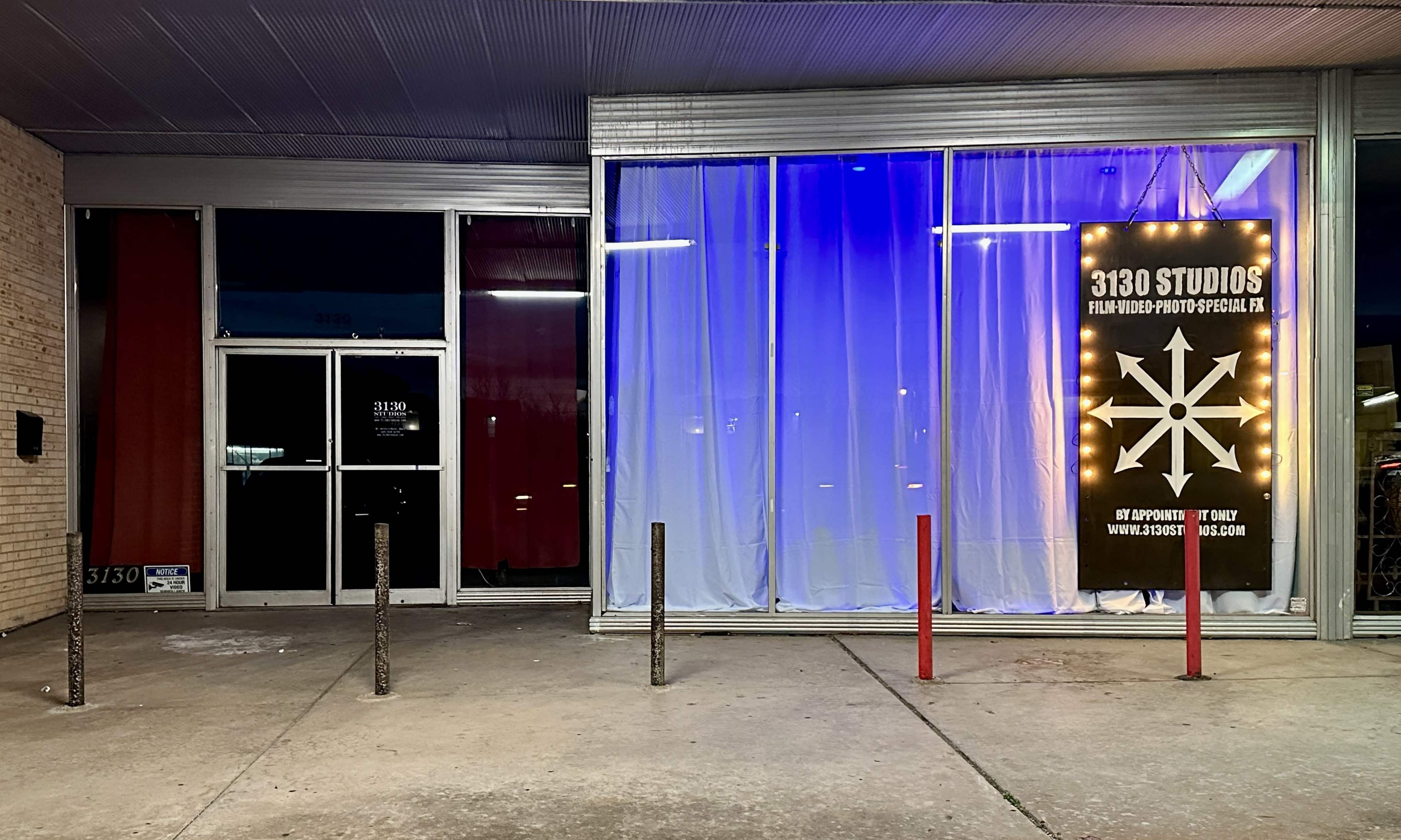The exterior of 3130 Studios features large windows draped with colorful curtains and a bright, illuminated sign beside the entrance.