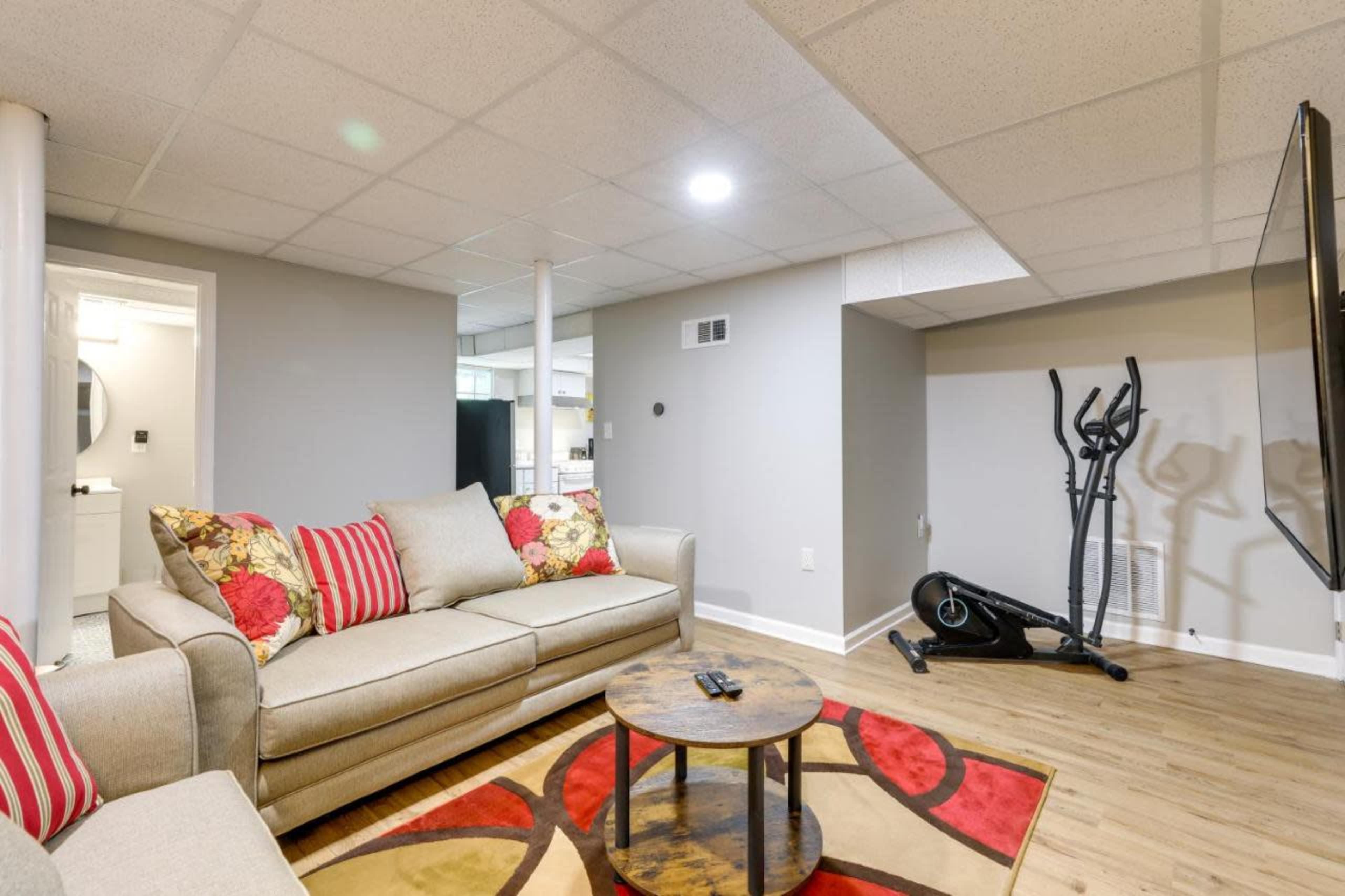 The image shows a finished basement featuring a beige couch with decorative pillows, a circular coffee table, a stationary exercise bike, and a flat-screen TV on the wall.