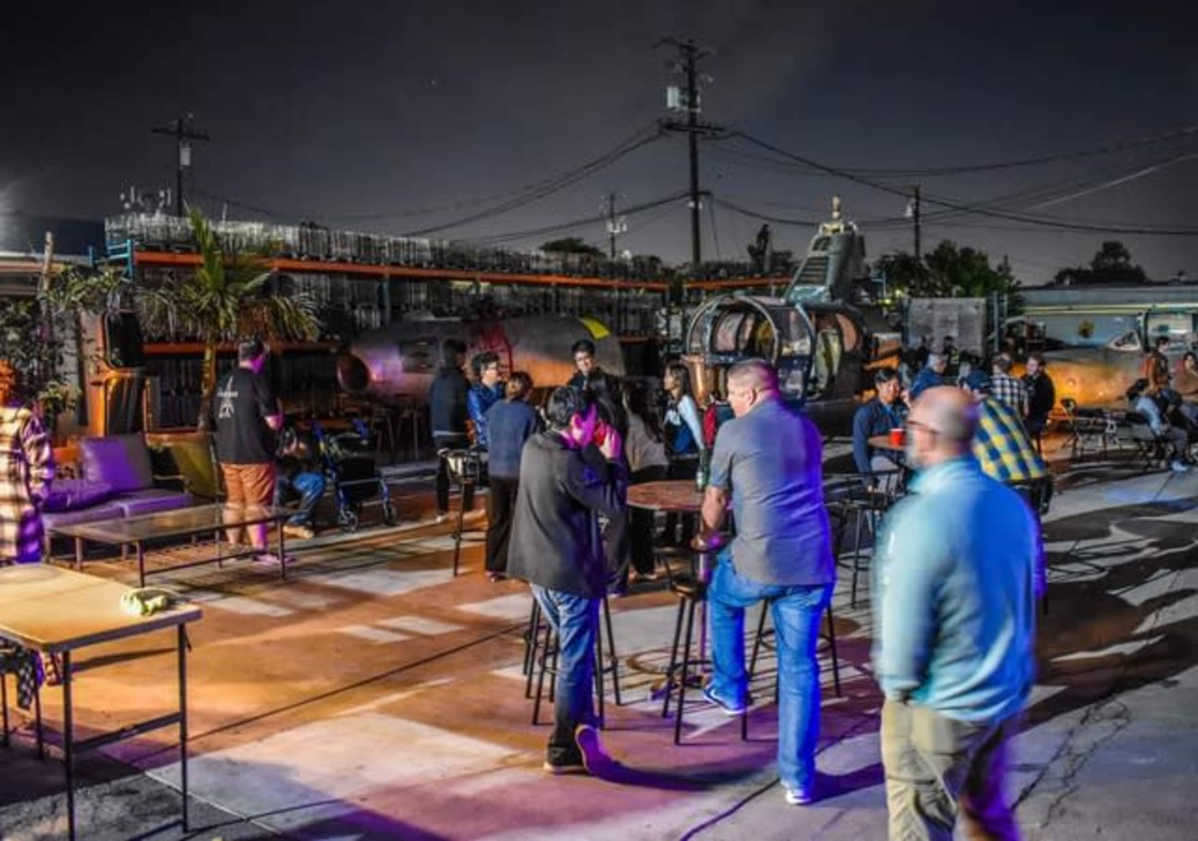 A lively outdoor gathering is taking place at night, featuring people socializing around tables amid industrial surroundings.