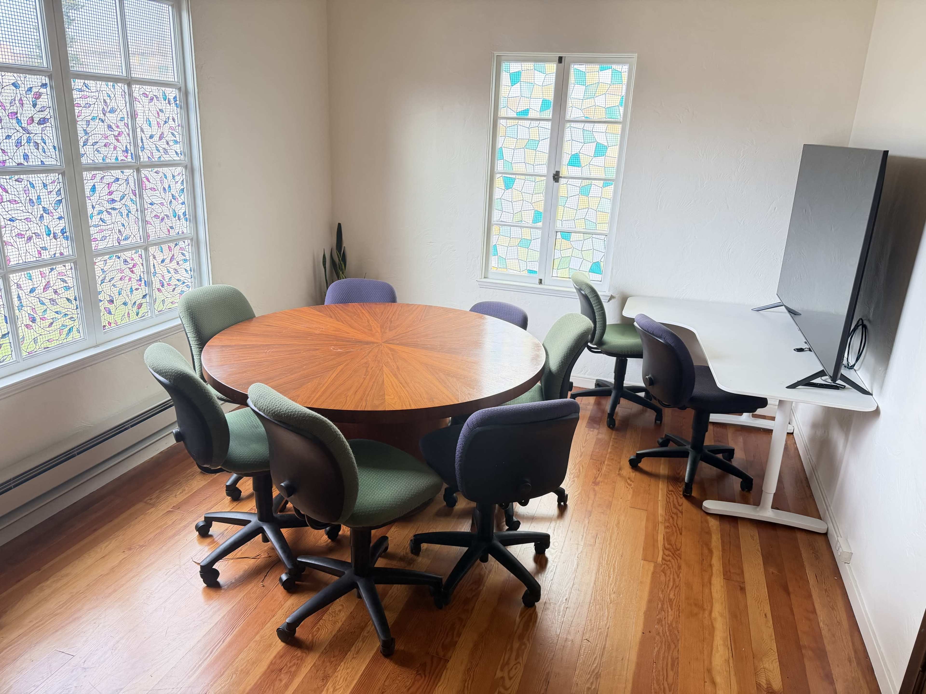The image shows a conference room with a circular wooden table surrounded by six ergonomic chairs and a wall-mounted television.