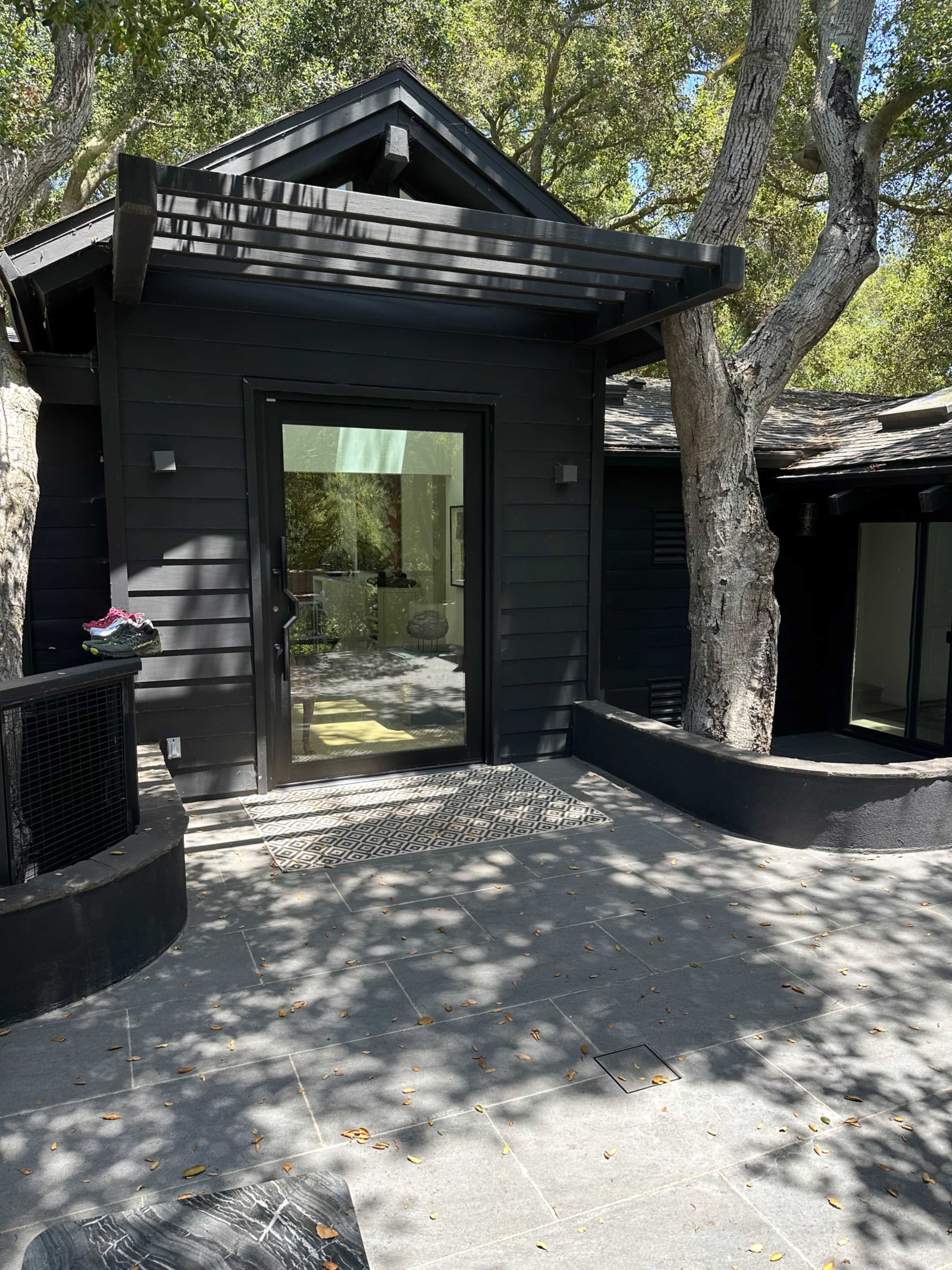 A dark-colored wooden house with a large glass door and a stone pathway surrounded by trees.