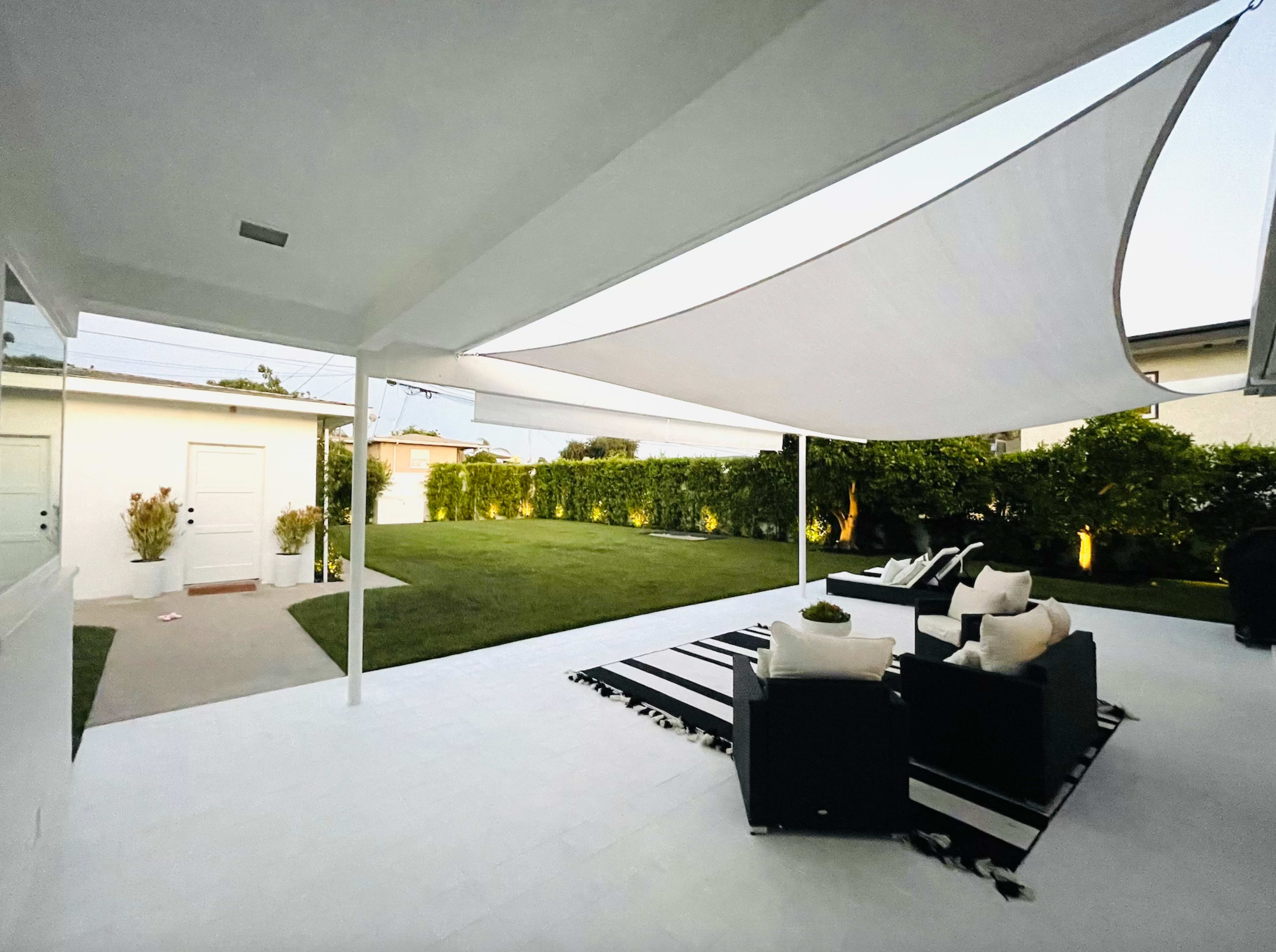 A modern outdoor living space features a shaded area with black seating, a striped rug, and a neatly manicured lawn surrounded by greenery.