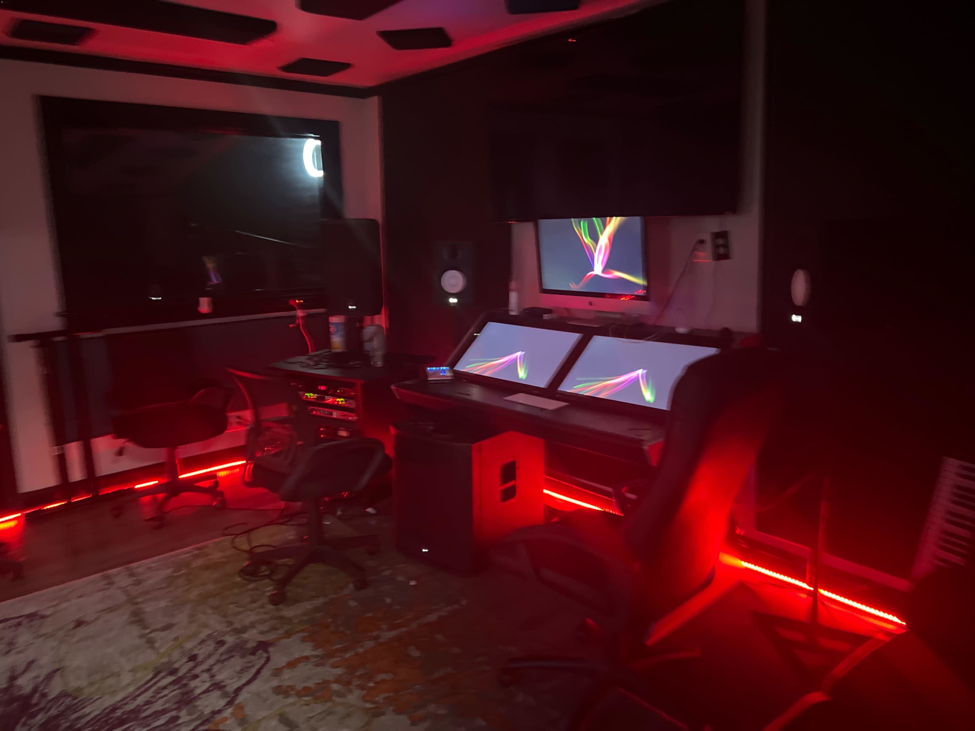 The image shows a dimly lit music studio featuring multiple screens, a mixing console, and several chairs arranged around a carpeted floor.
