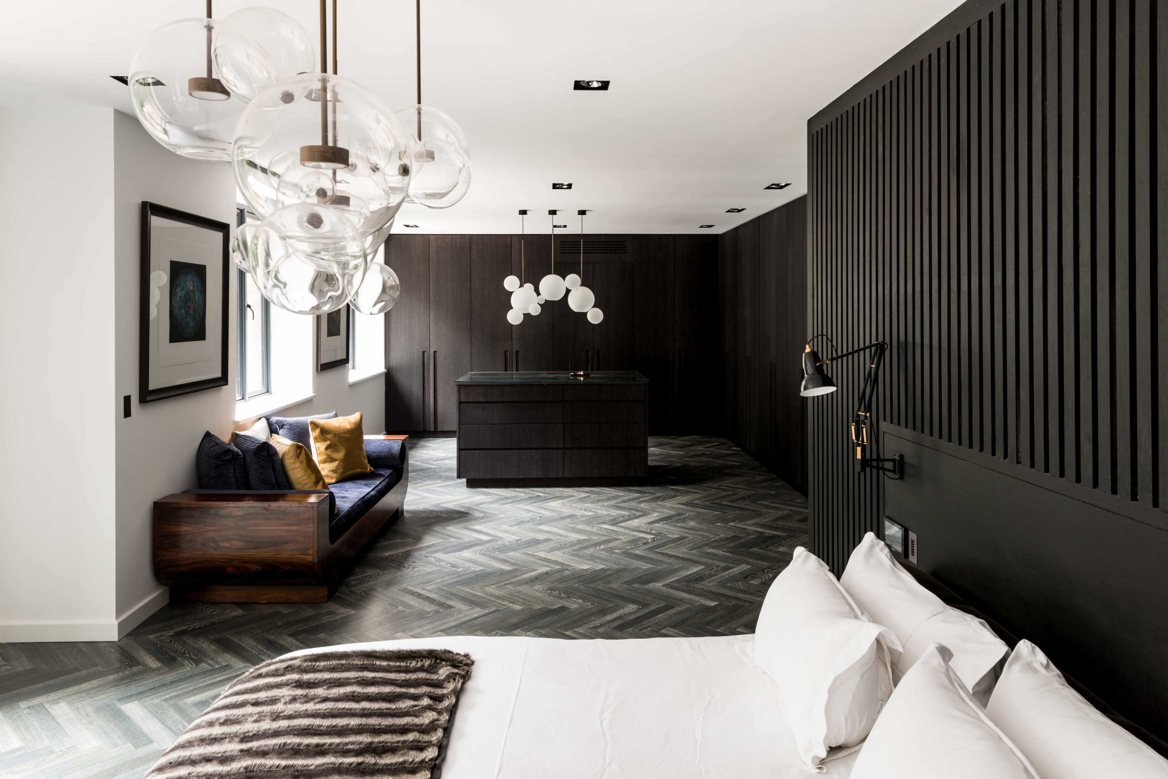 The image shows a modern bedroom with a large bed, a minimalist seating area, and a stylish kitchen island, all featuring a mix of dark wood and light colors.