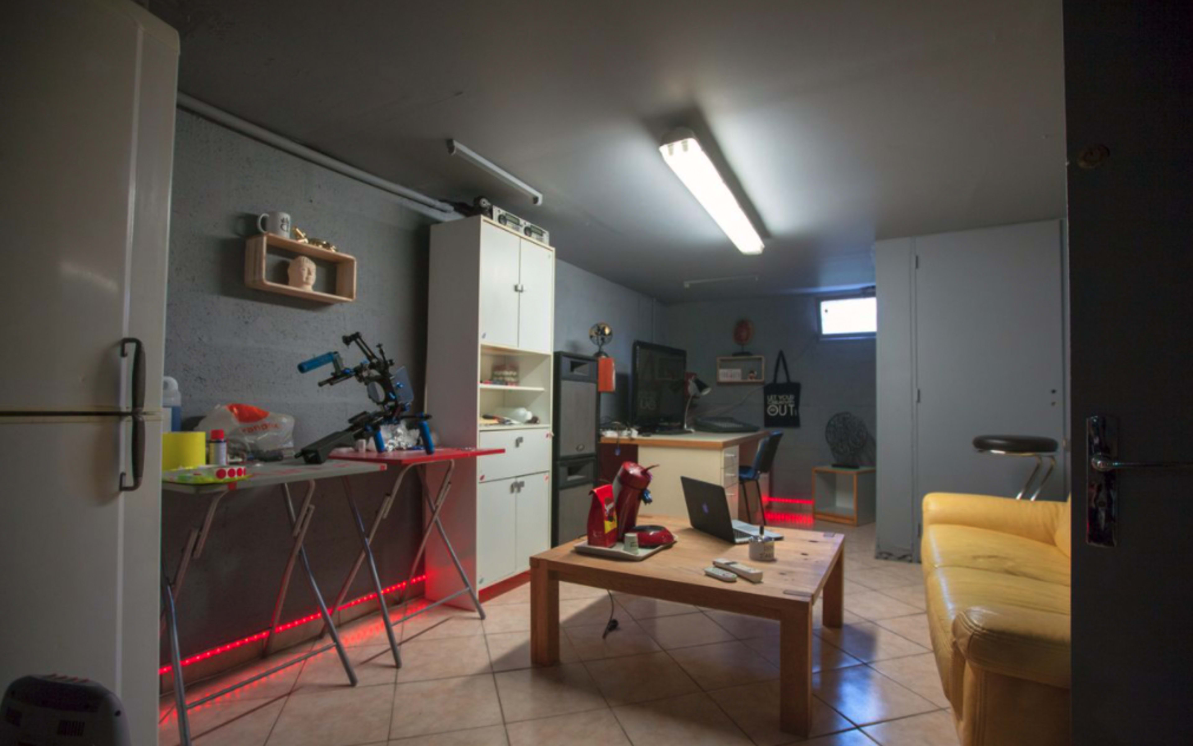 A modern basement room featuring a kitchen area with a refrigerator, a desk with a laptop, and a small seating area with a yellow couch.