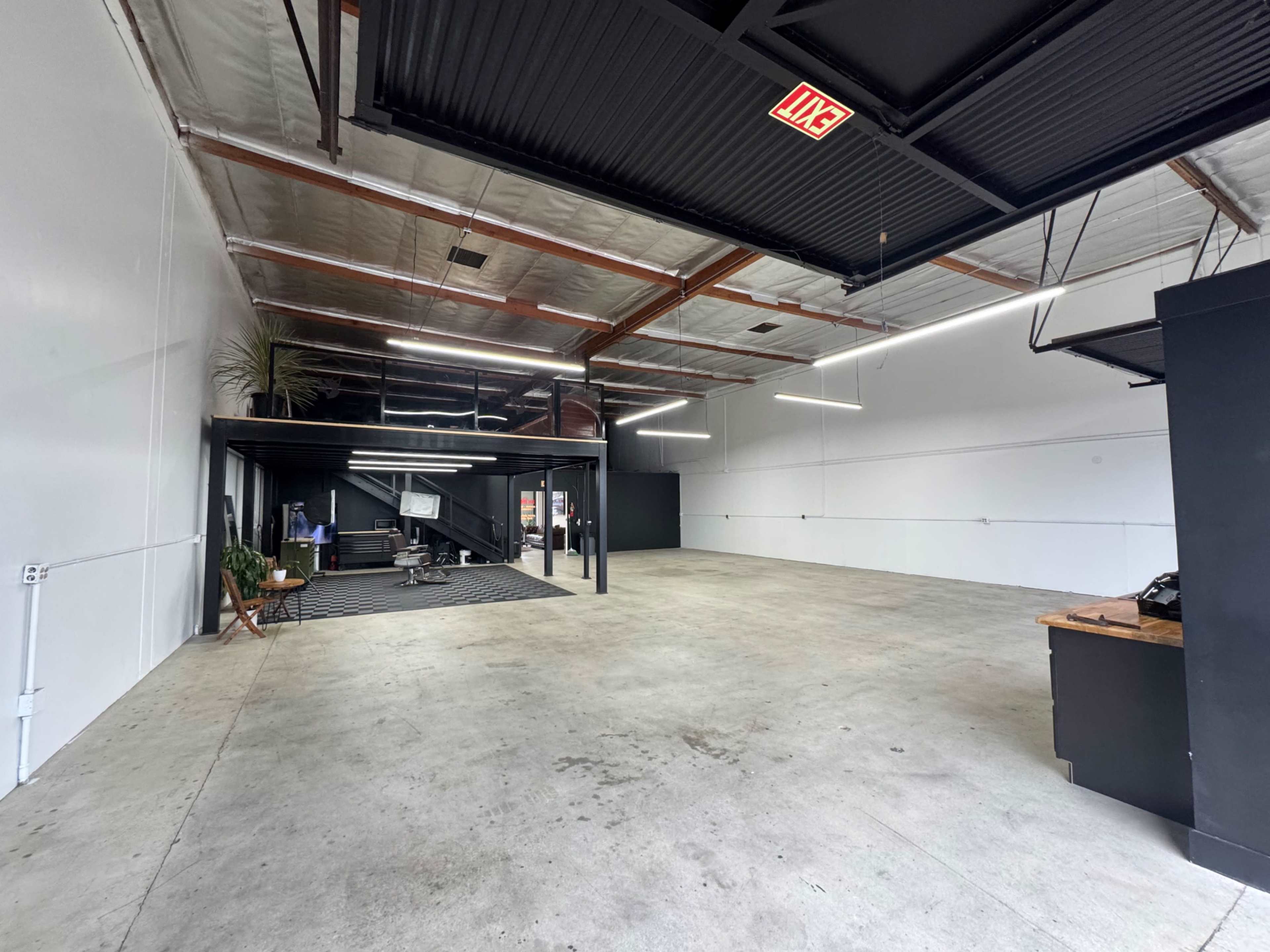 The image shows a spacious, empty warehouse with concrete flooring, exposed beams, and a sectioned area featuring a staircase and a dark-finished counter.
