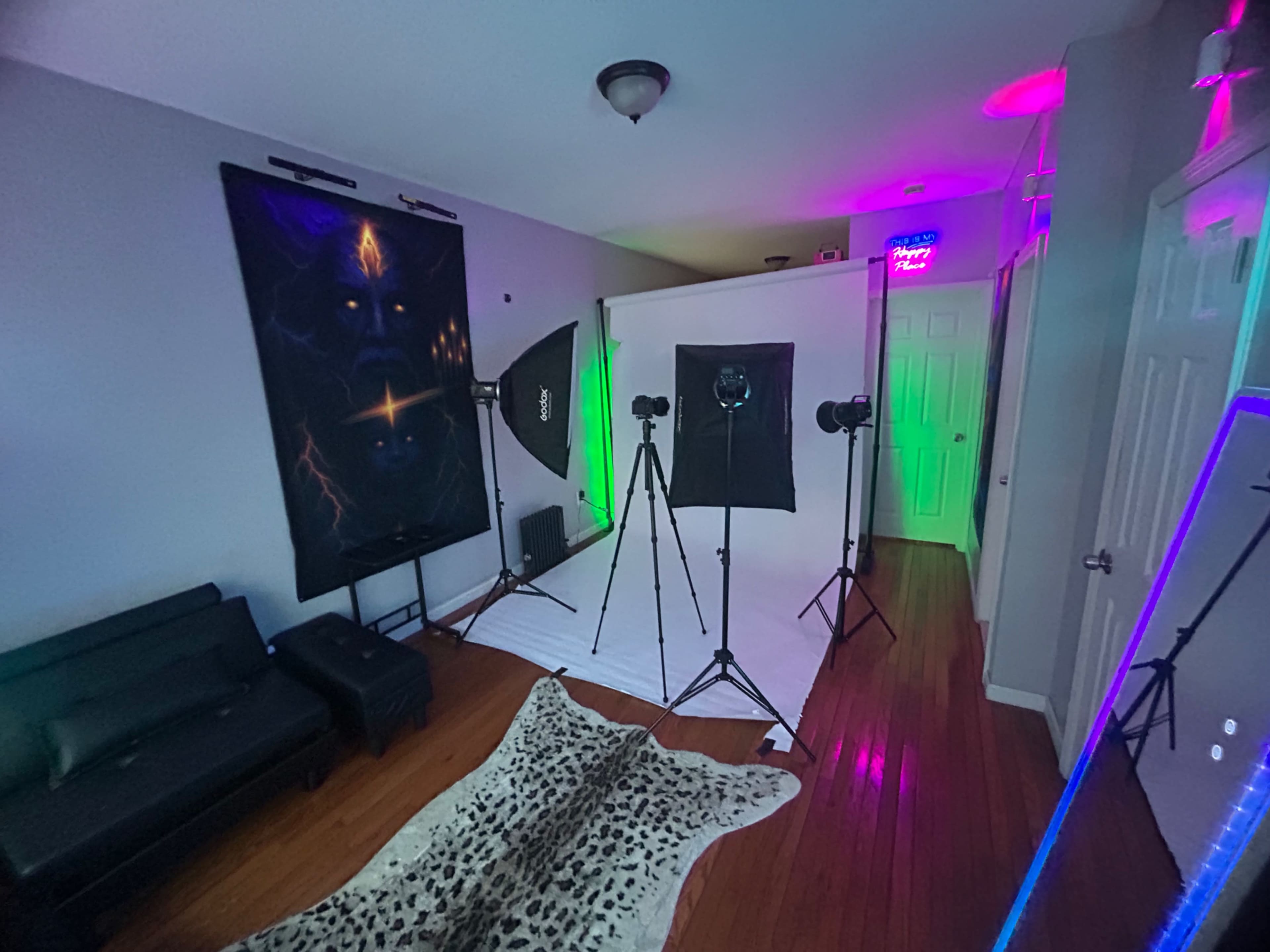 The image shows a photo studio setup with a white backdrop, two tripods with cameras, and colorful lighting illuminating the walls.