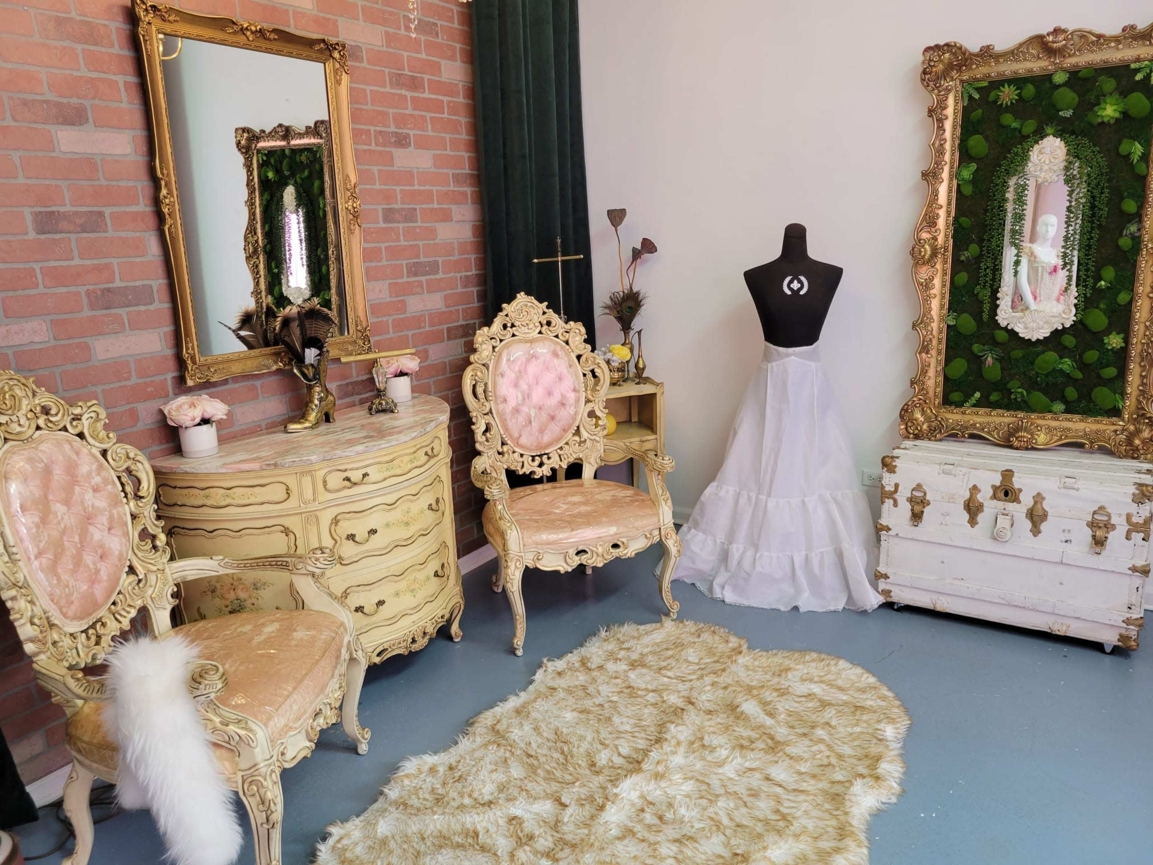 The interior features a vintage-style room with ornate golden furniture, including two chairs and a dresser, alongside a dress form and a decorative mirror surrounded by greenery.