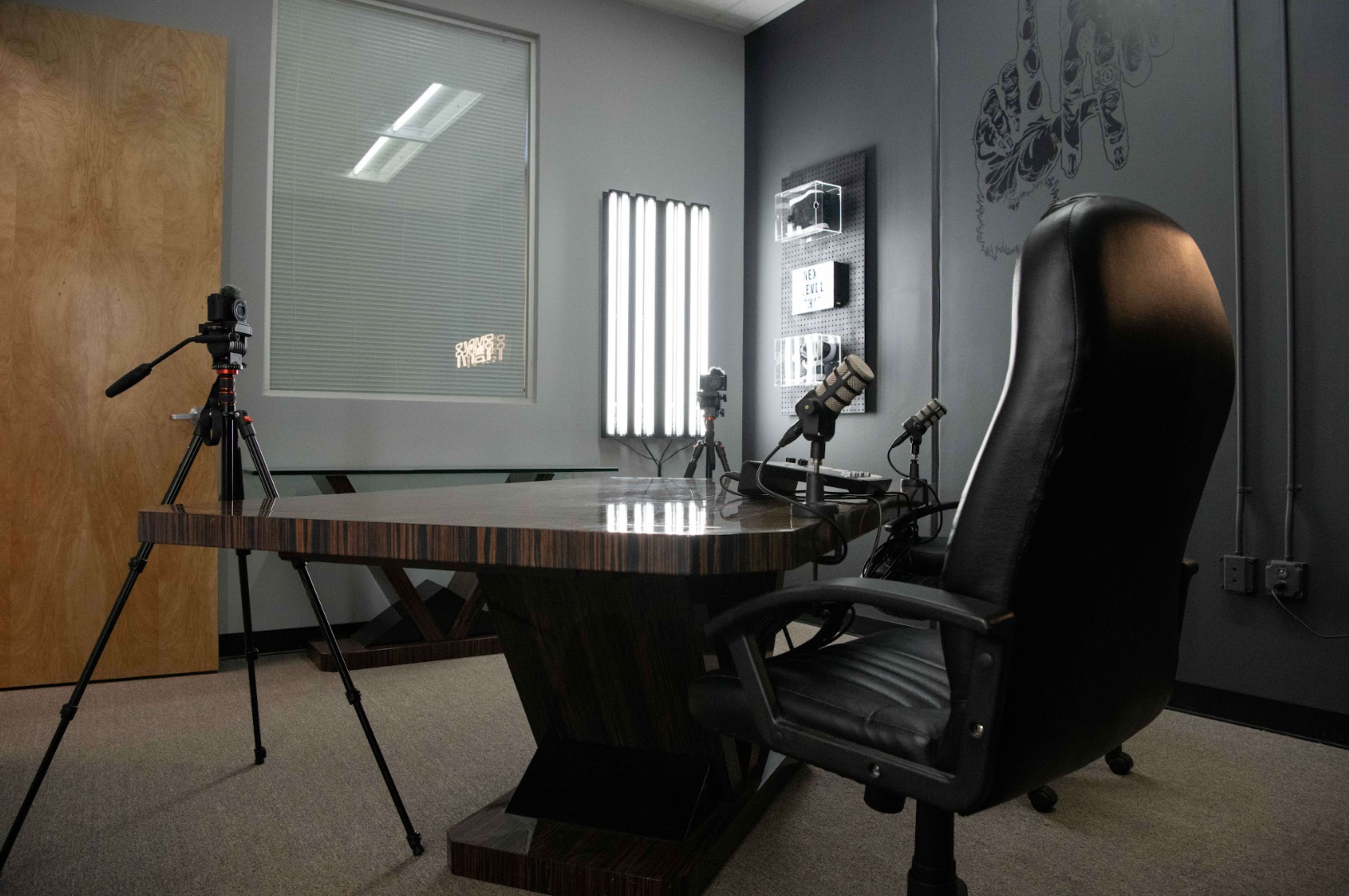 A modern office space featuring a large dark wooden table, a high-backed black chair, and two tripods with cameras set up on either side.