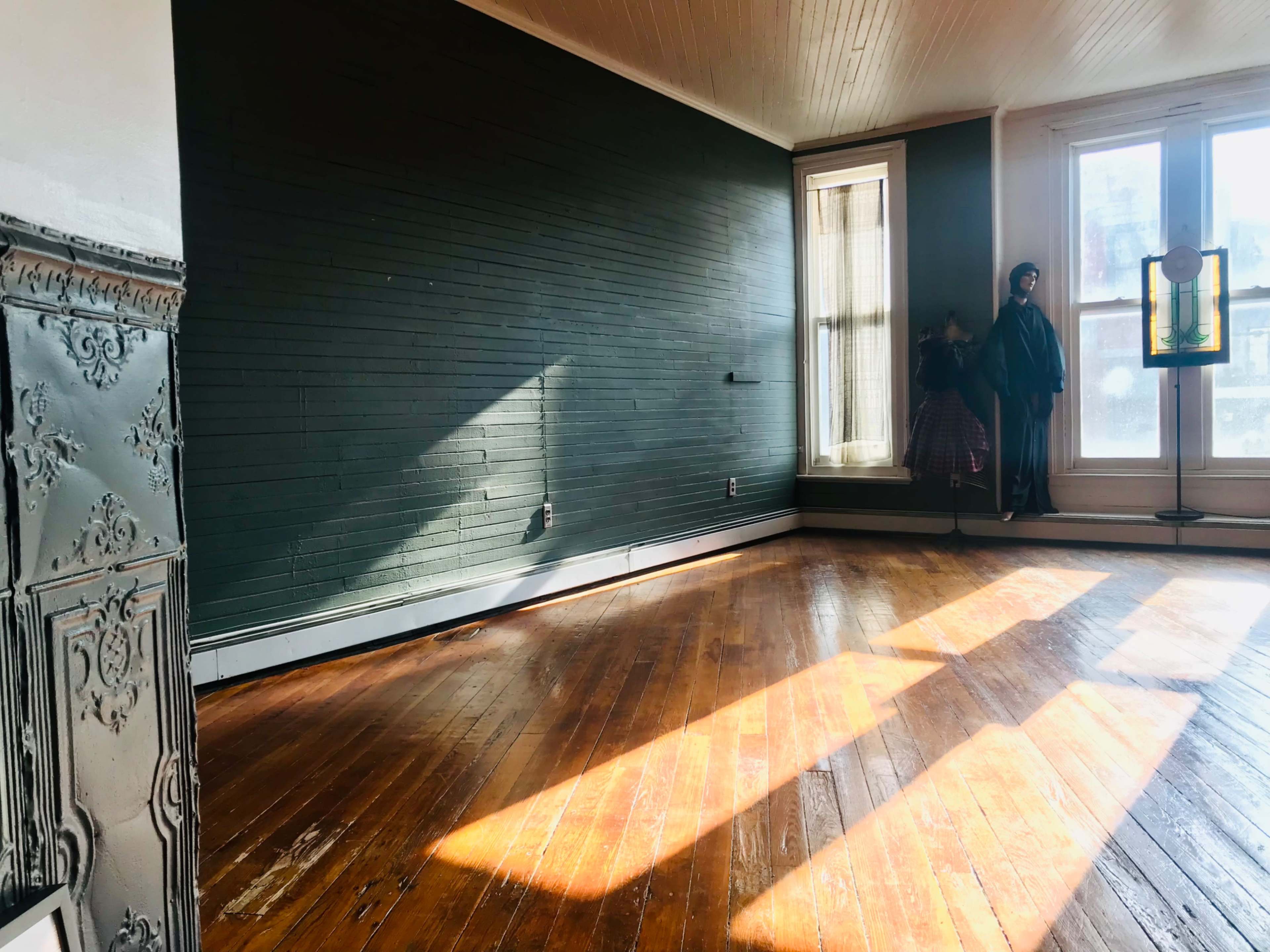 The image shows a sunlit room with wooden floors, a dark green wall, and a tall window casting long shadows across the space.