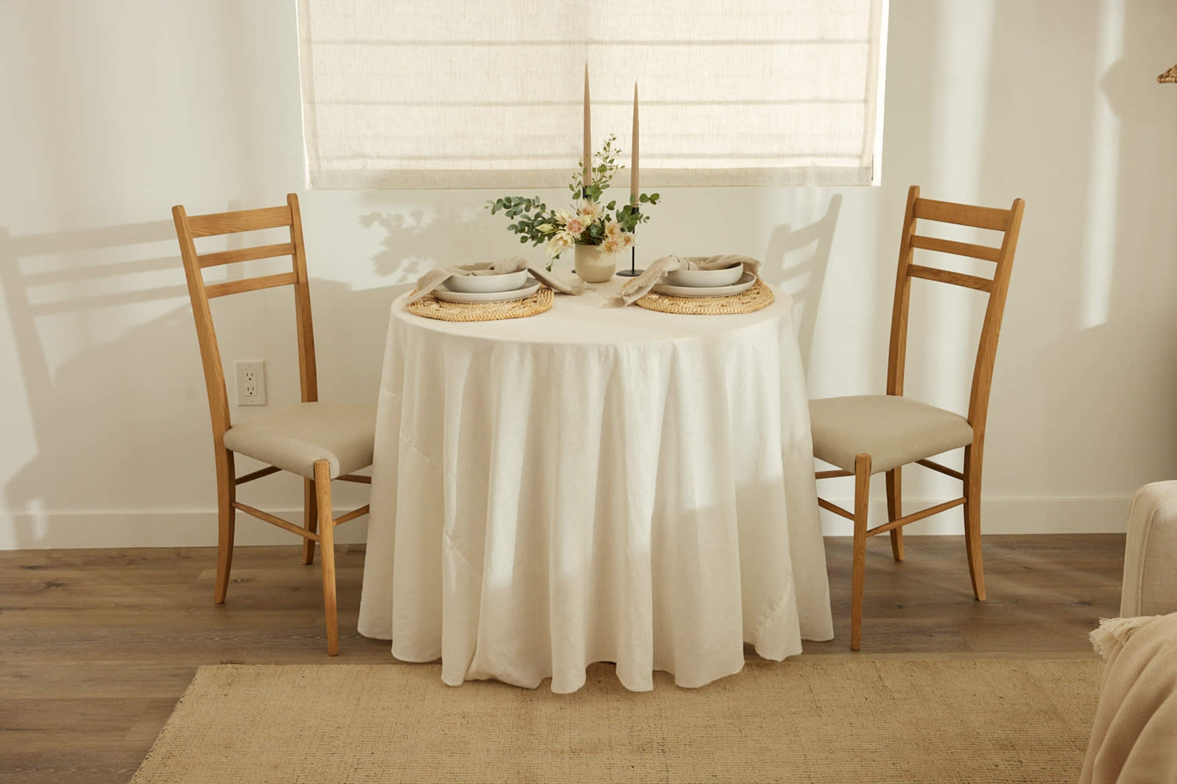 A round table covered with a white tablecloth is set for two with plates and a small flower arrangement, accompanied by two wooden chairs.