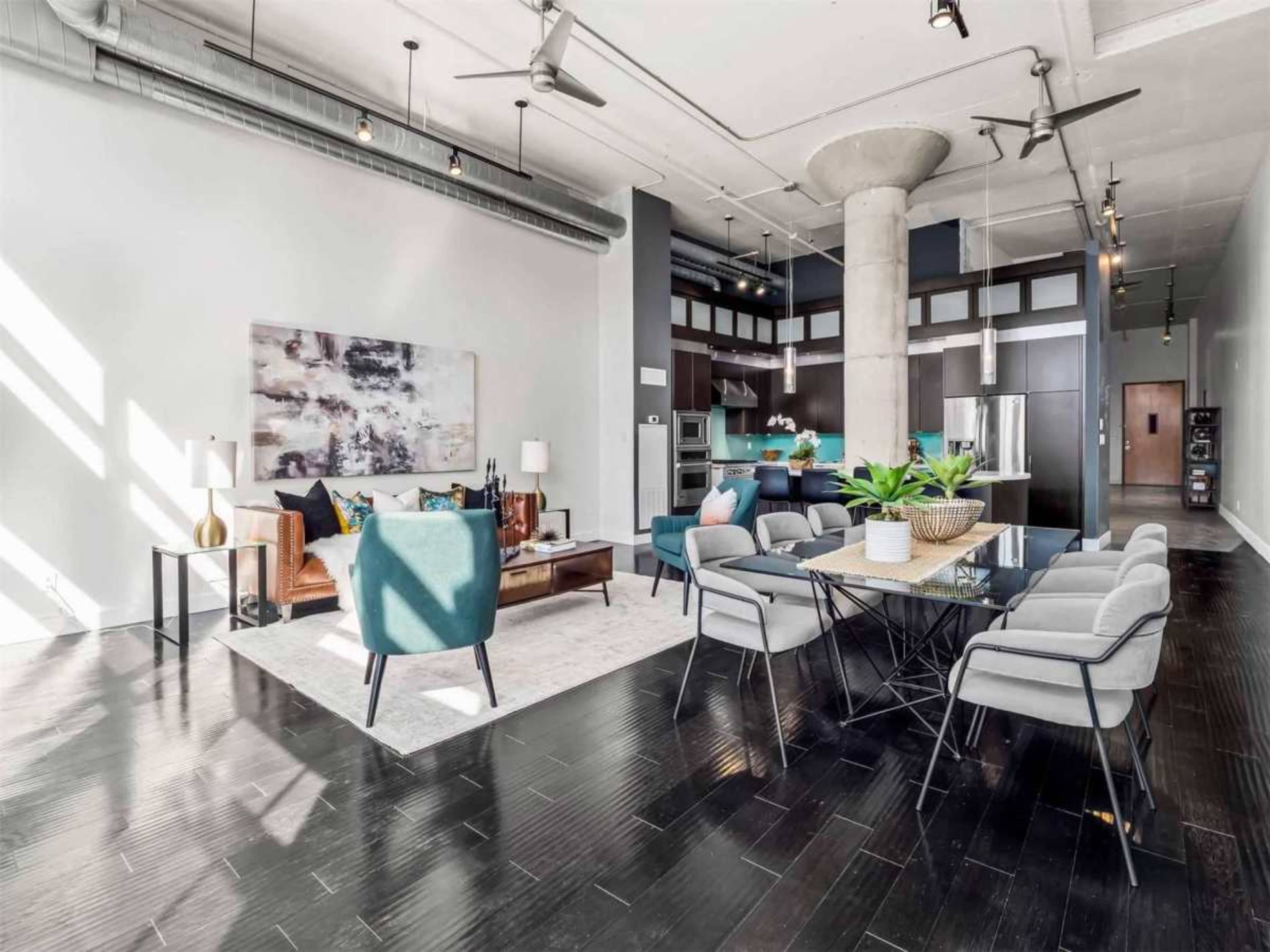 The image shows a modern, spacious living area with a dining table, comfortable seating, and large windows allowing natural light to enter.