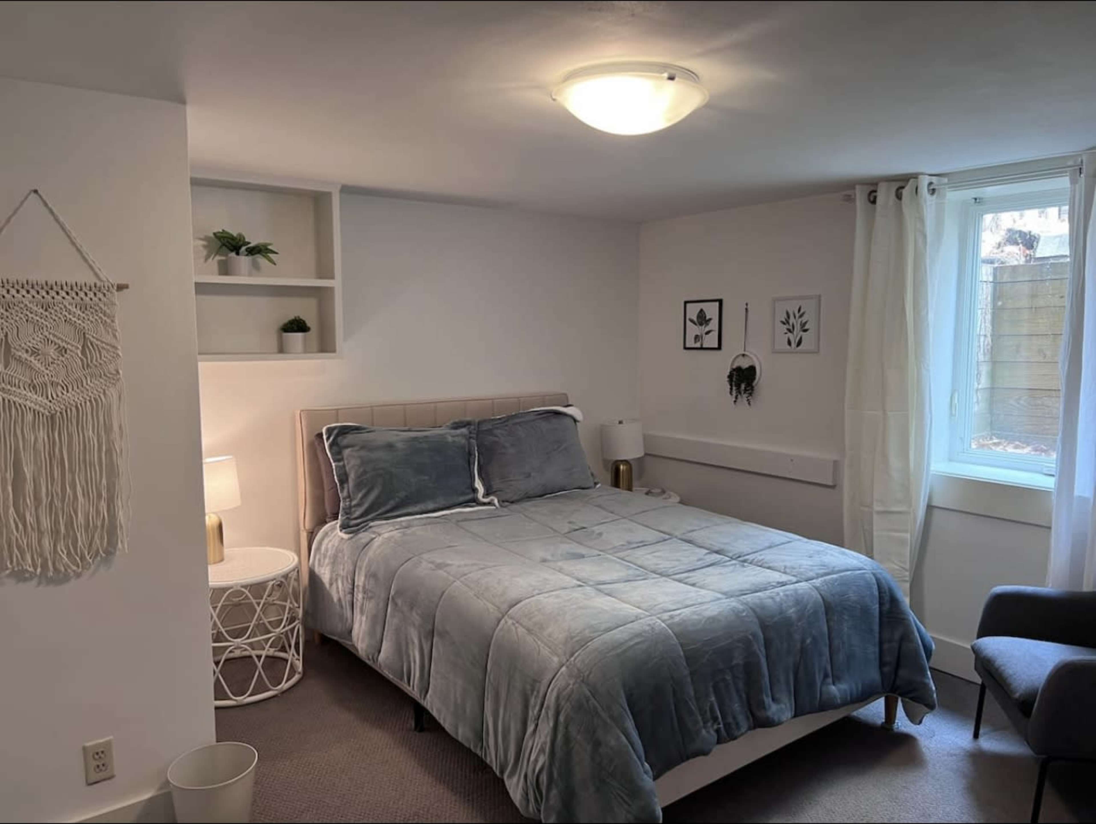 A softly lit bedroom features a bed with a light blue blanket, a nightstand with a lamp, and decorative wall art.