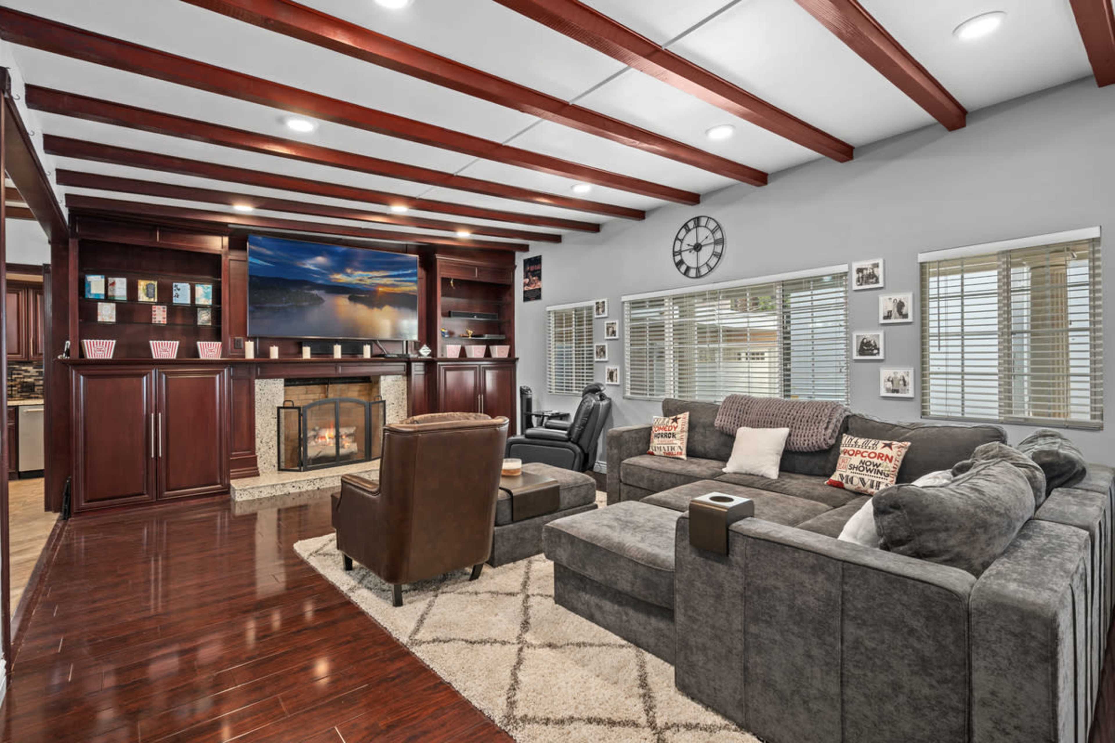 The living room features a sectional sofa, two armchairs, a wall-mounted TV above a fireplace, and wood beams on the ceiling, with large windows providing natural light.