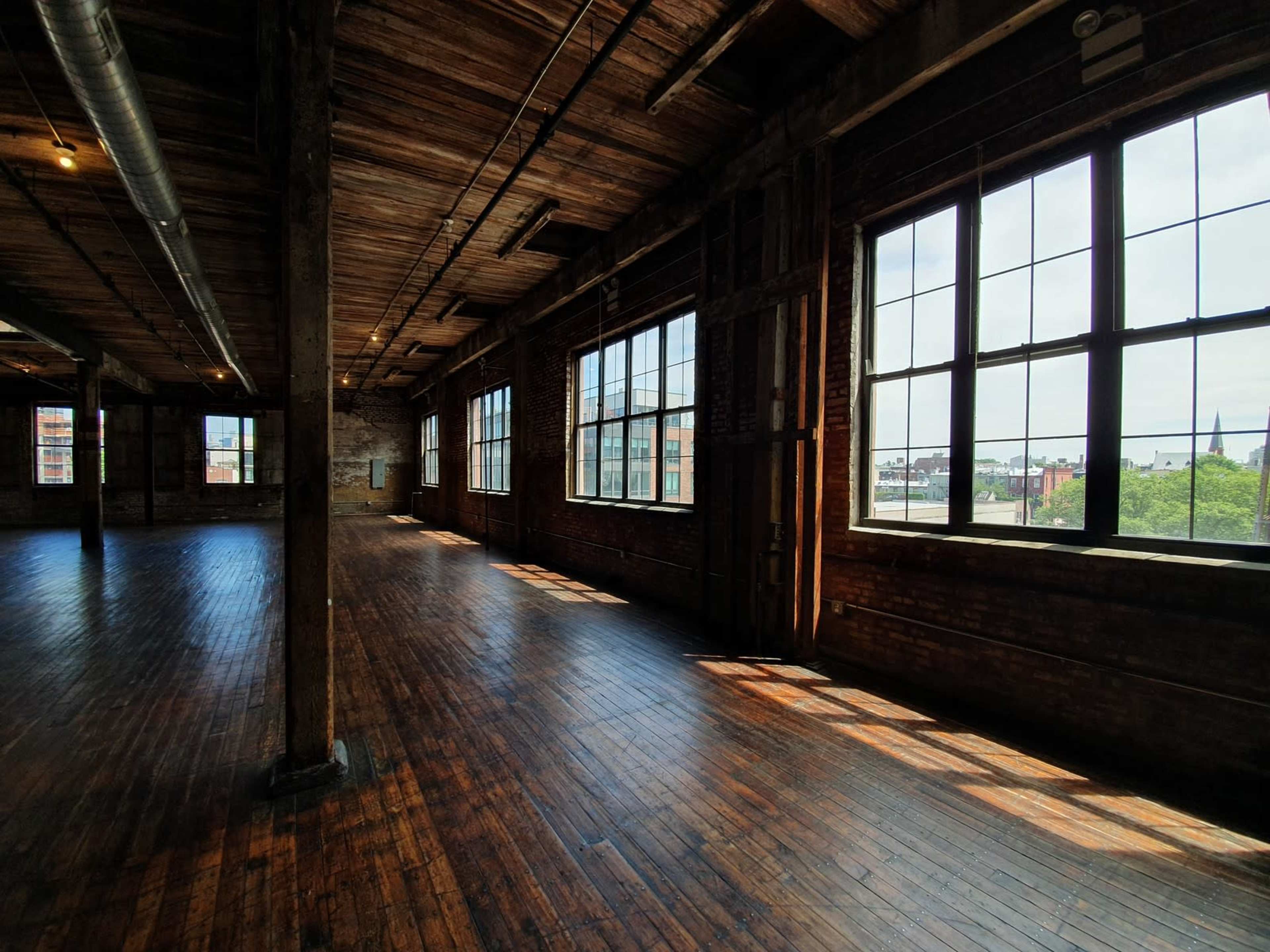 A spacious, industrial room with large windows and exposed wooden beams, featuring polished wooden floors and ample natural light.