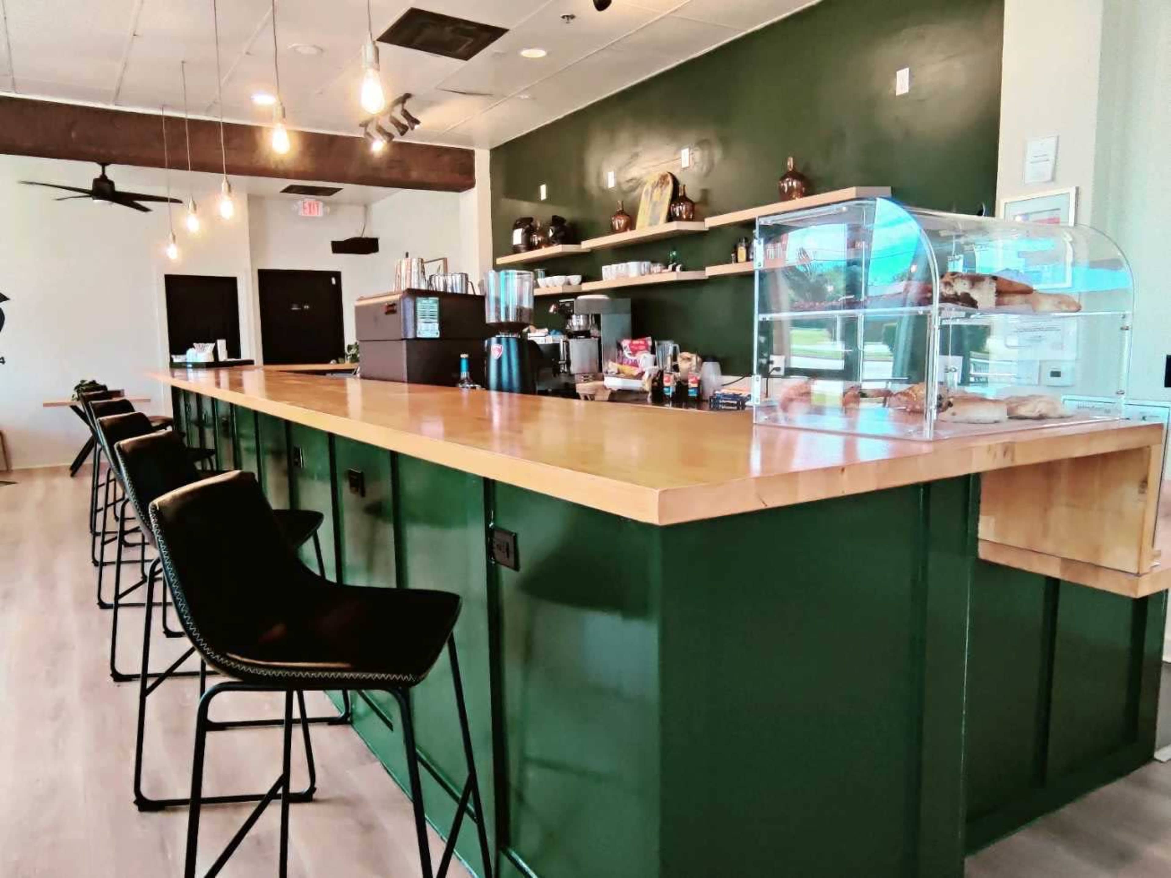 The image shows a modern café interior with a green bar counter, high stools, and a display case for baked goods.