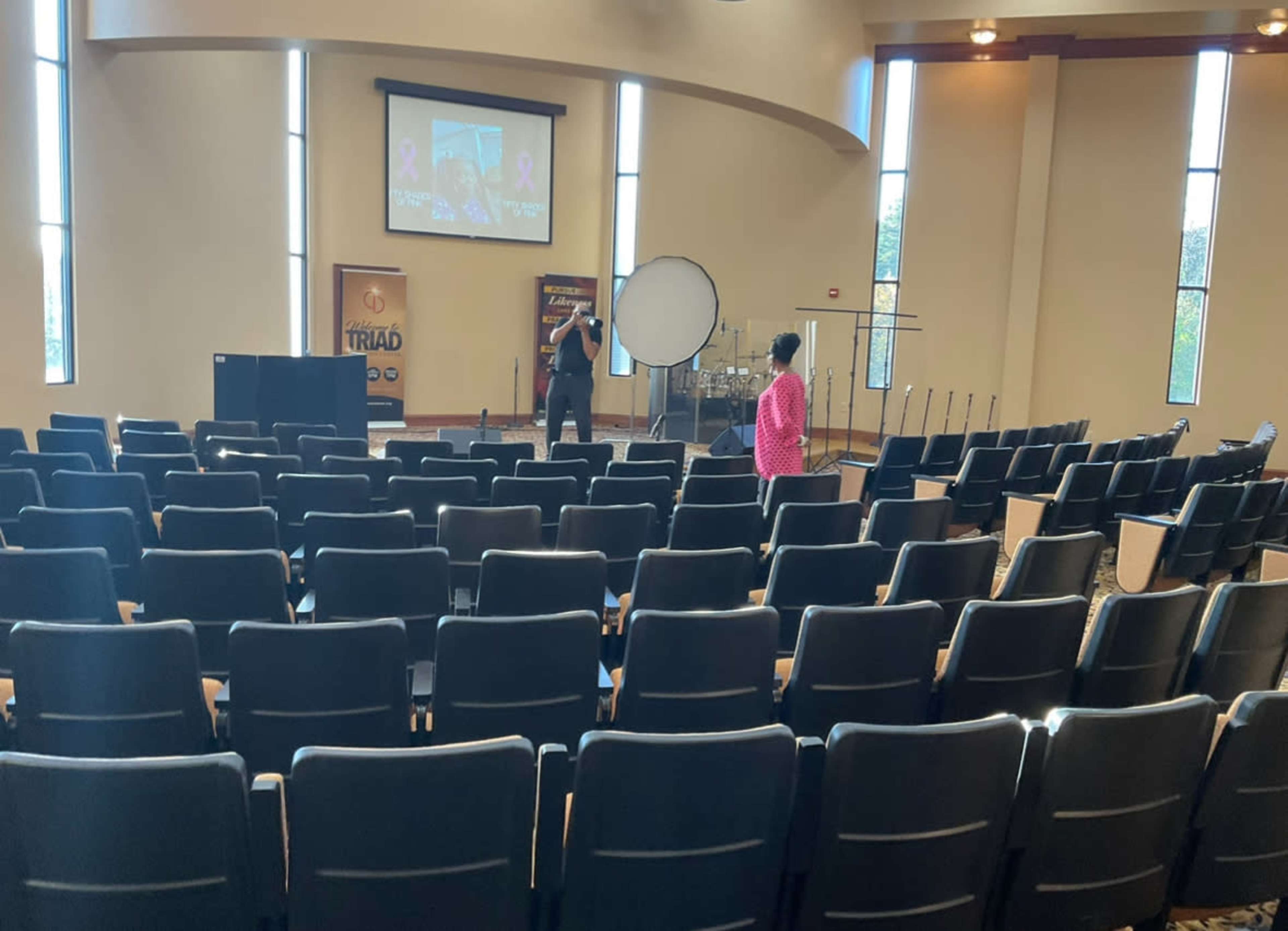 A man is standing on a stage in a mostly empty auditorium, preparing for a presentation while a woman observes from the front row.