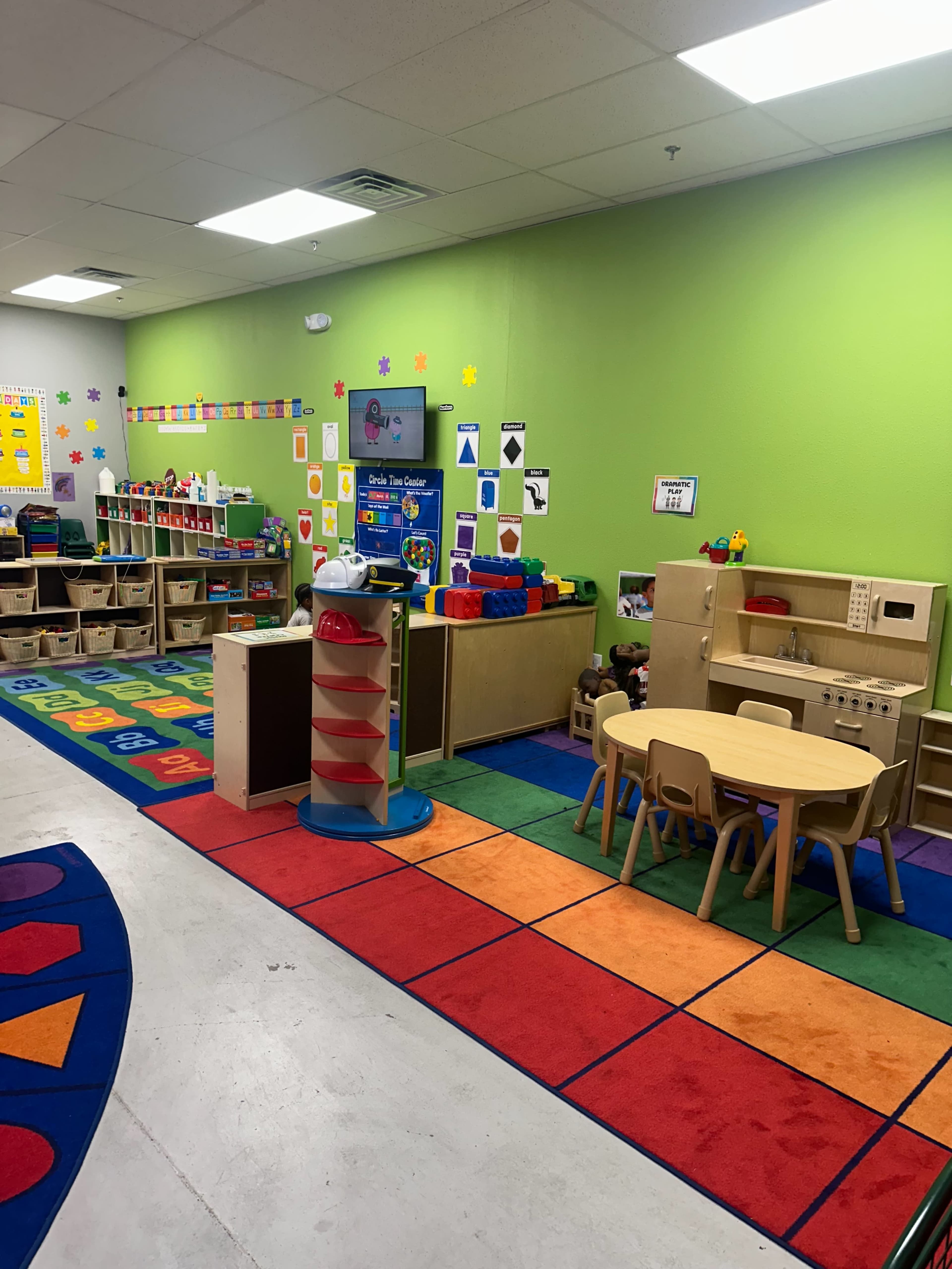 The image shows a colorful preschool classroom with shelves of toys, a circular table with chairs, and bright green walls.