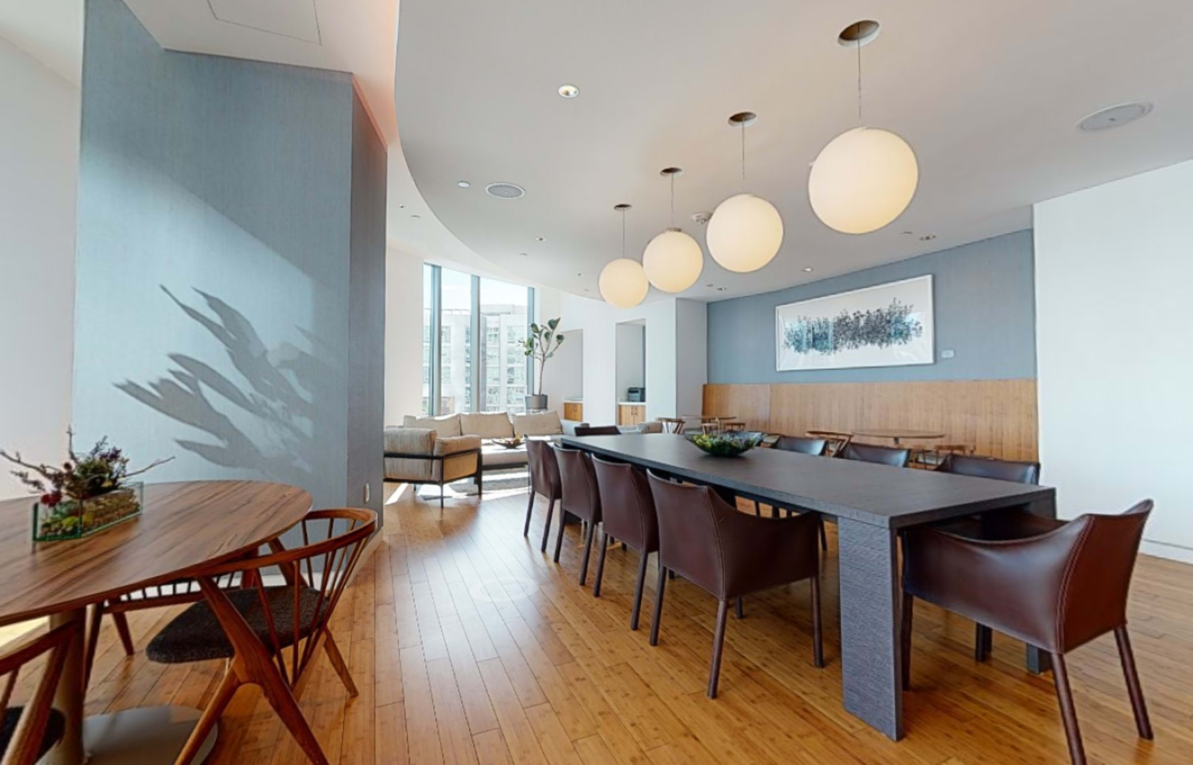 A modern dining area featuring a long table with chairs, illuminated by round pendant lights, and surrounded by large windows.