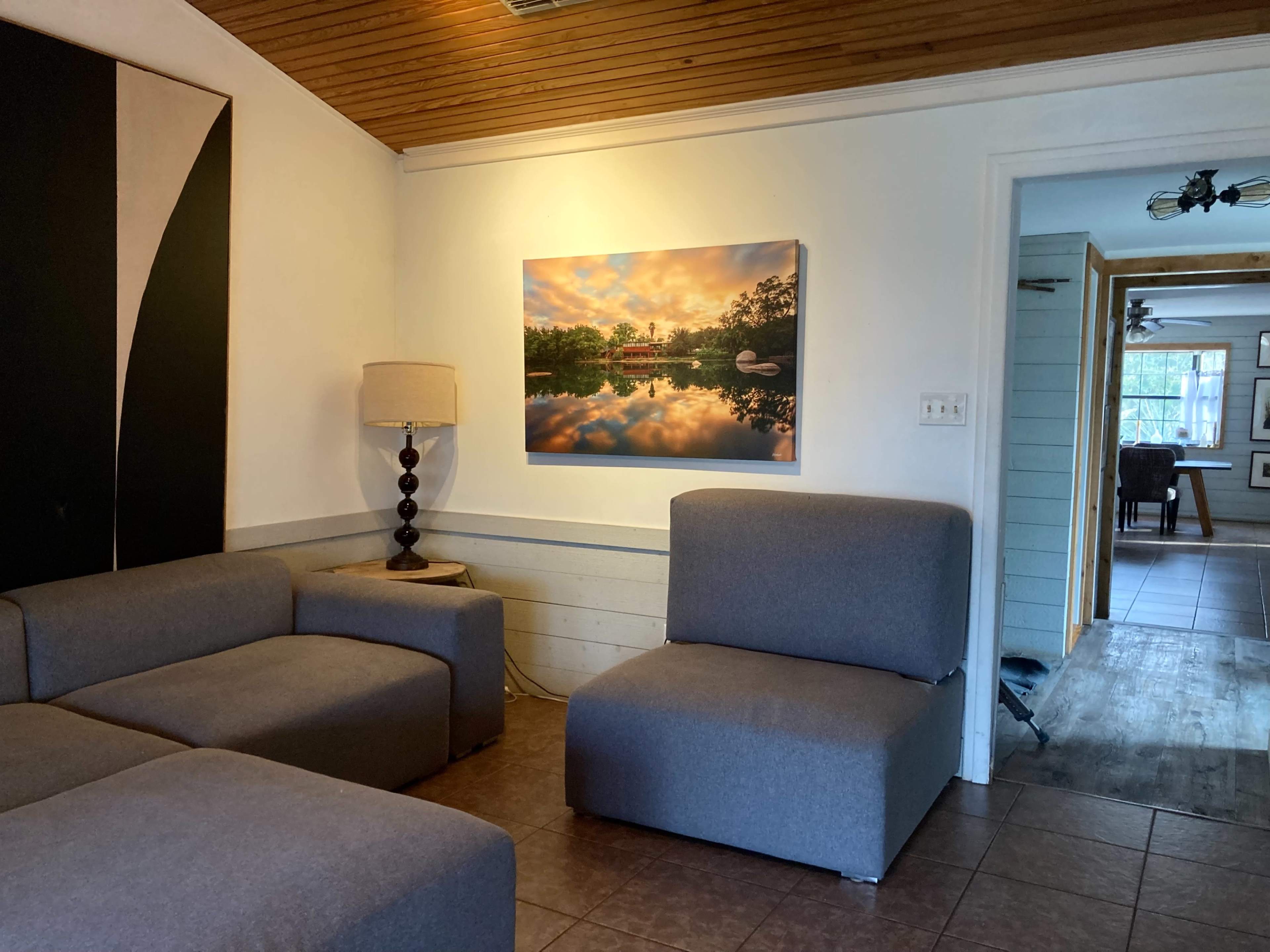 A living room with gray modular seating, a lamp, and a large landscape painting of a lake with trees, mounted on the wall.