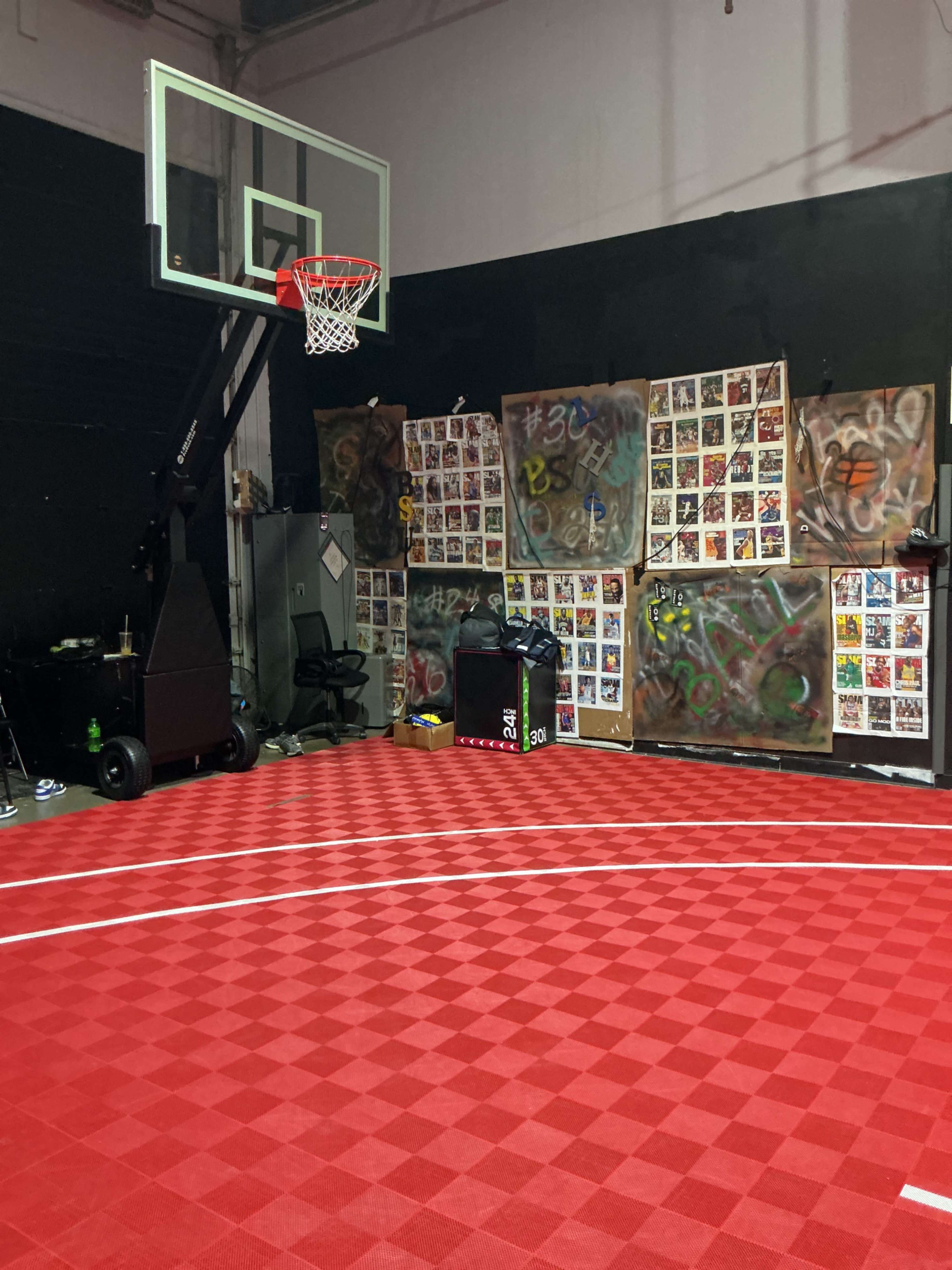 The image shows a gymnasium with a red patterned basketball court, a hoop, and walls decorated with colorful posters and graffiti.