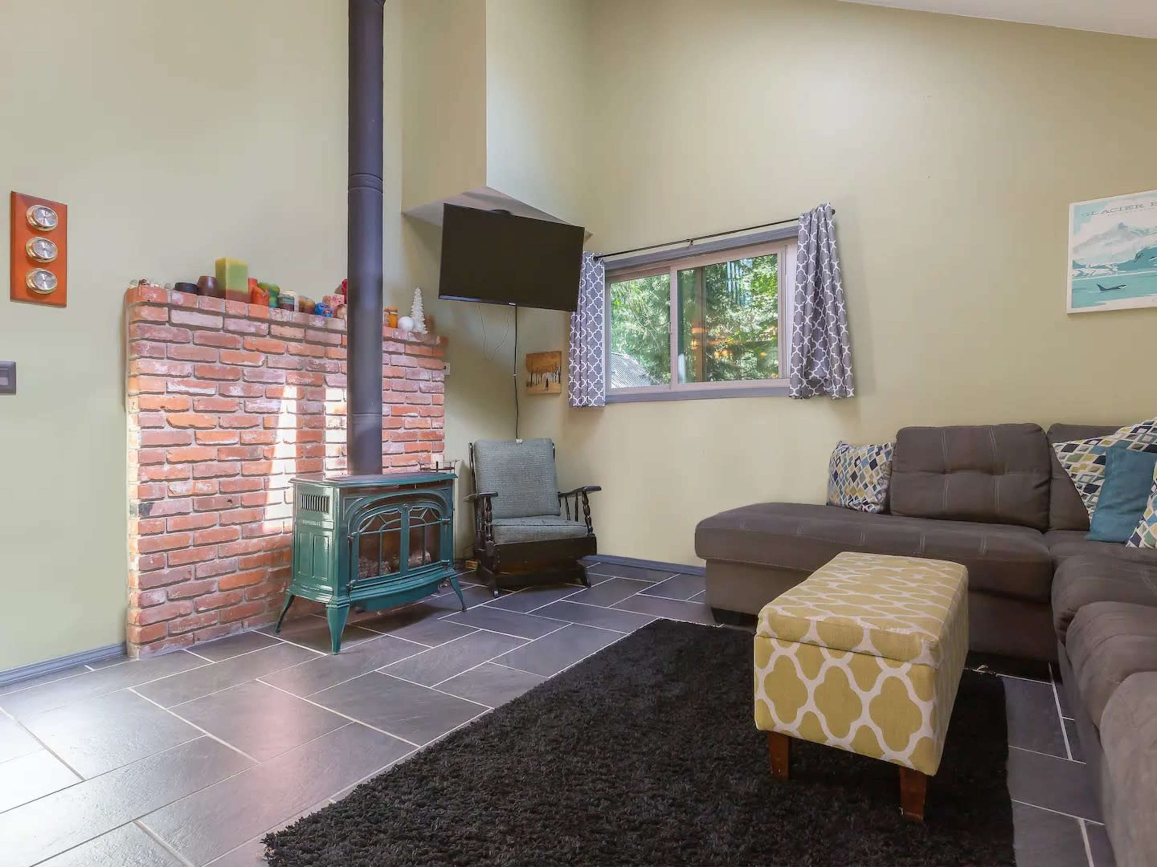 The room features a brick fireplace with a green stove, a large sofa, and a window overlooking trees.