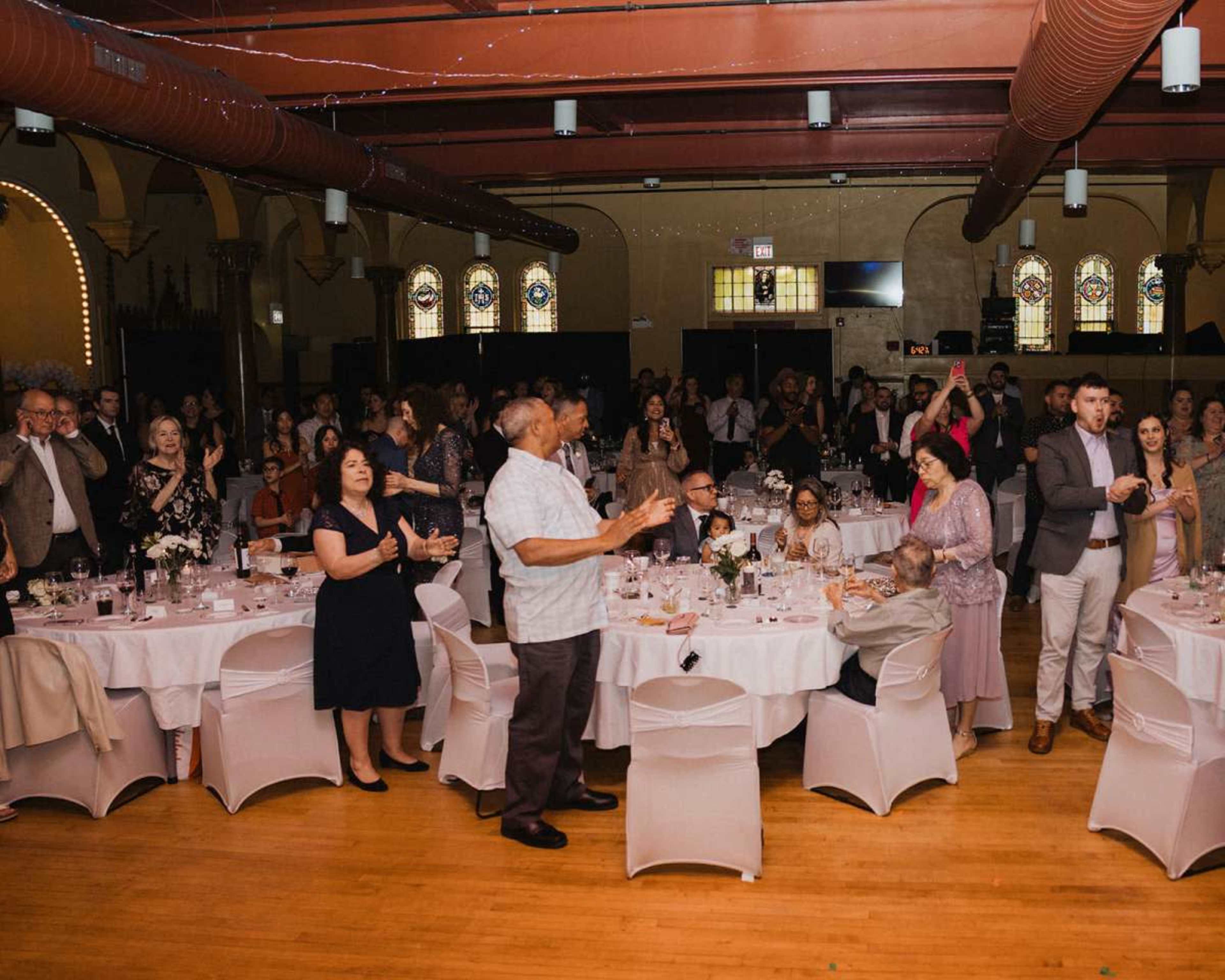 A crowd of guests stands and applauds at a formal event held in a spacious hall with decorative stained glass windows.