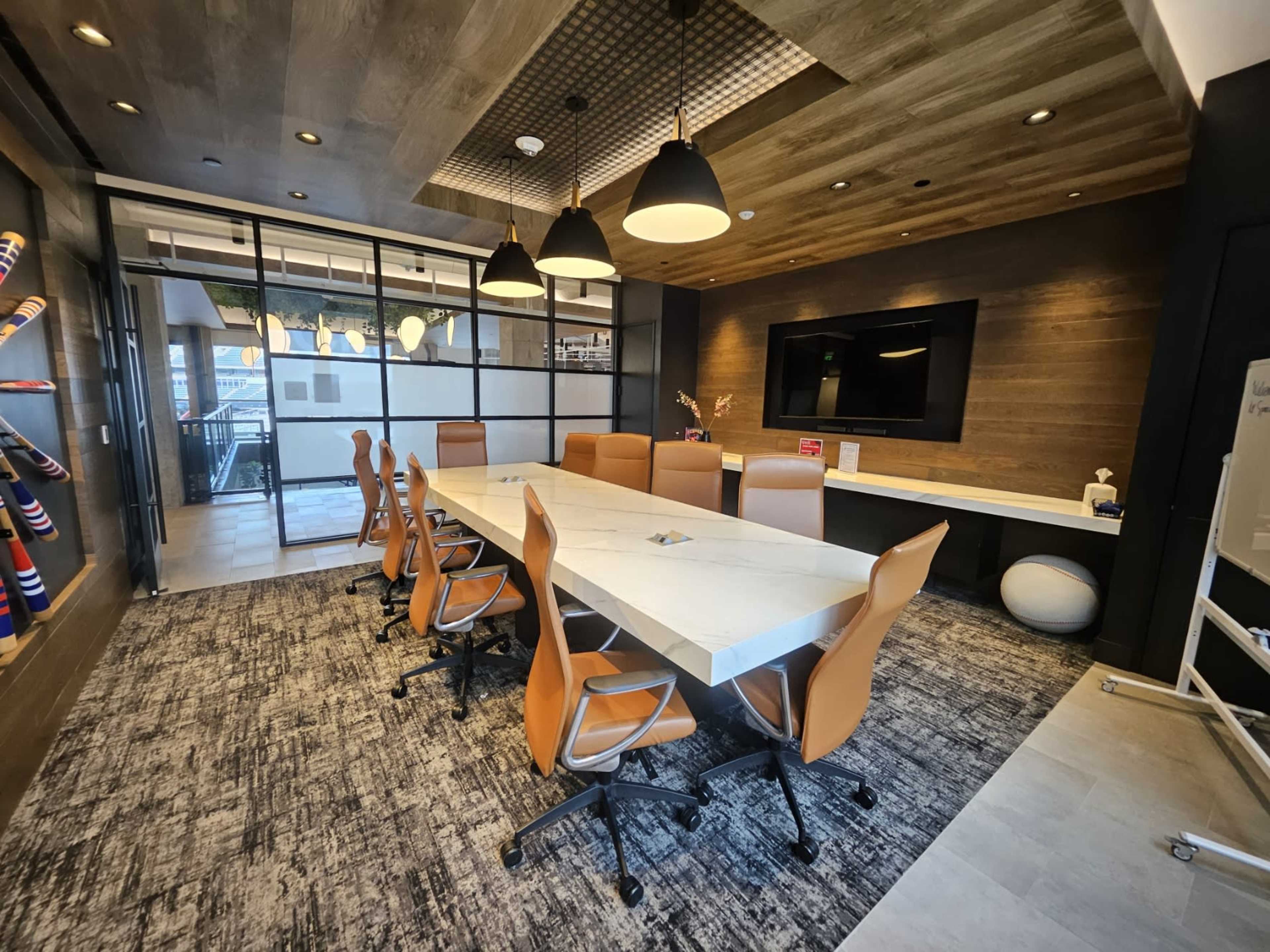 The image shows a modern conference room with a long white table surrounded by orange chairs, featuring wooden accents and a television on the wall.