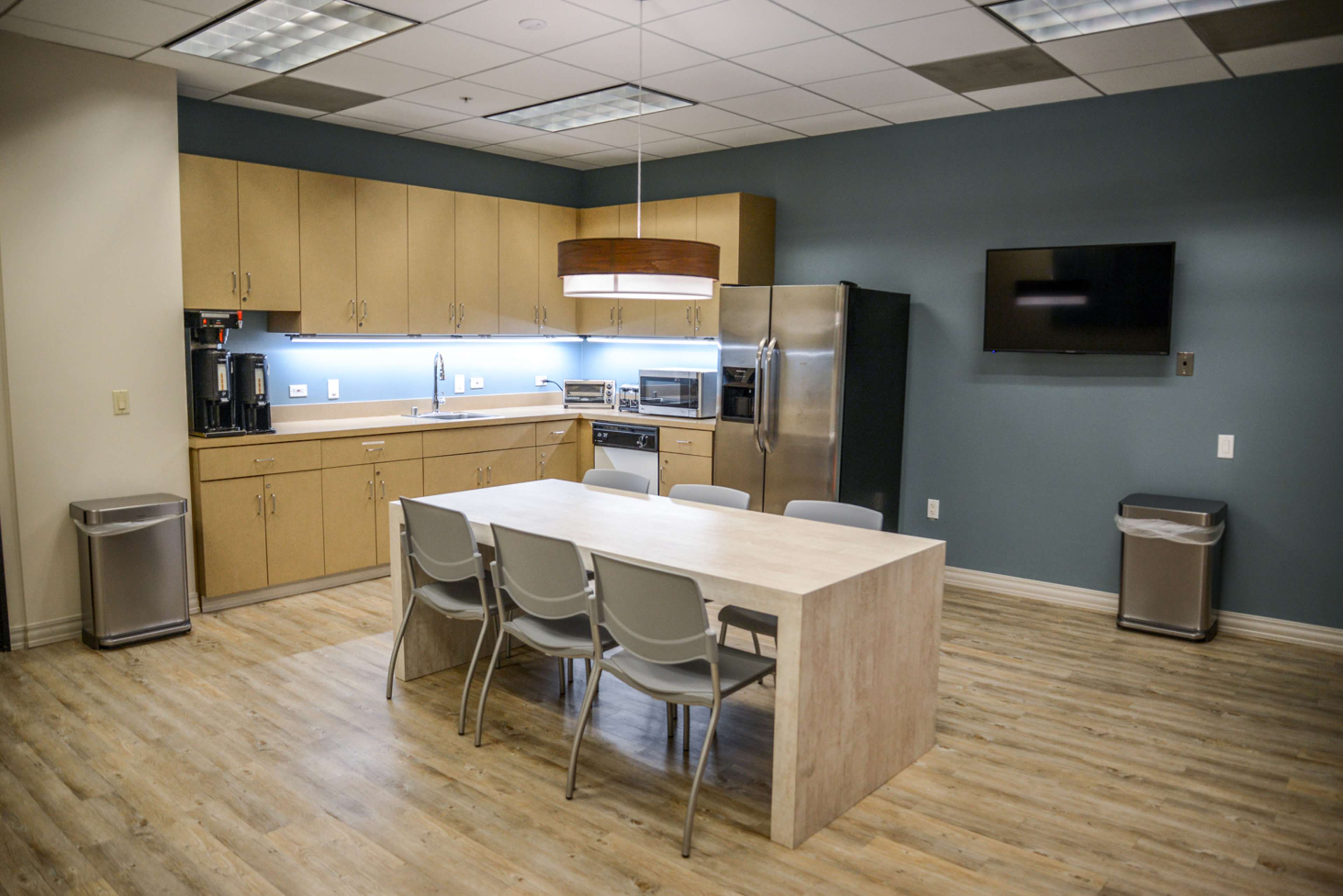 A modern kitchen break room with a table and chairs, stainless steel appliances, and a flat-screen TV mounted on the wall.