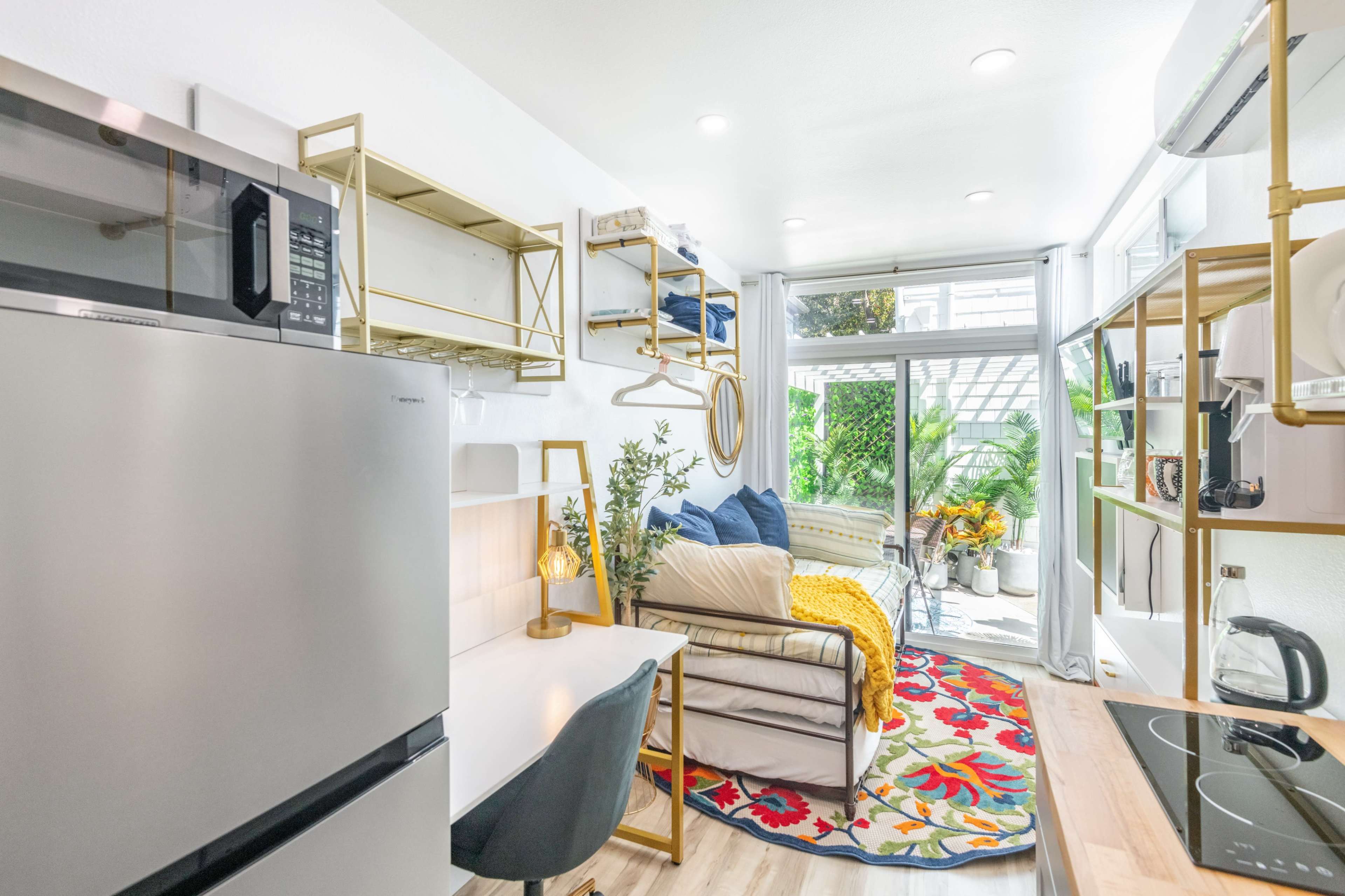 The image depicts a compact living space featuring a small kitchen area with a refrigerator and stove, a seating area with a couch, and a window that lets in natural light.