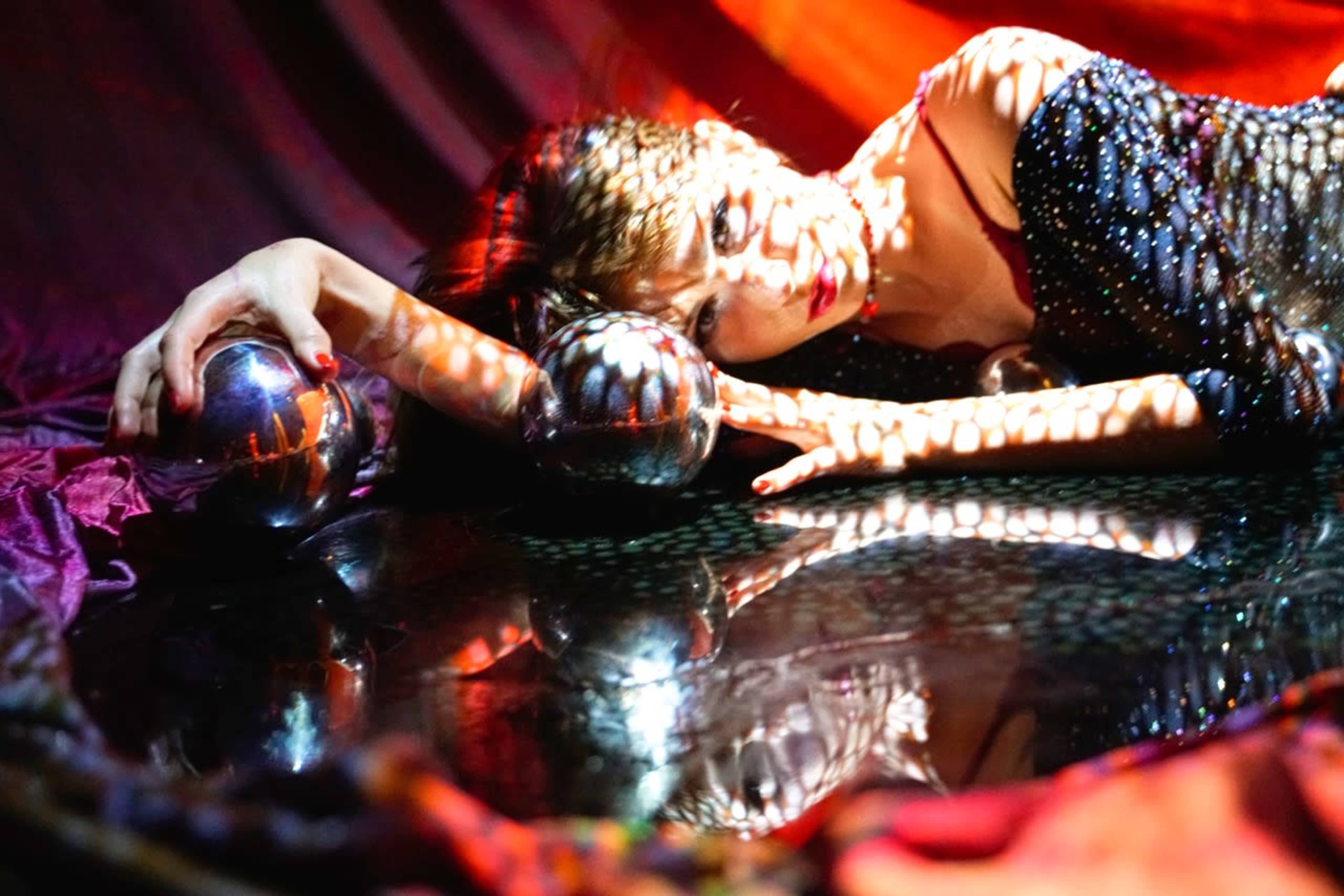 A woman lies on a reflective surface surrounded by silver spheres, with patterned shadows cast across her face and body.