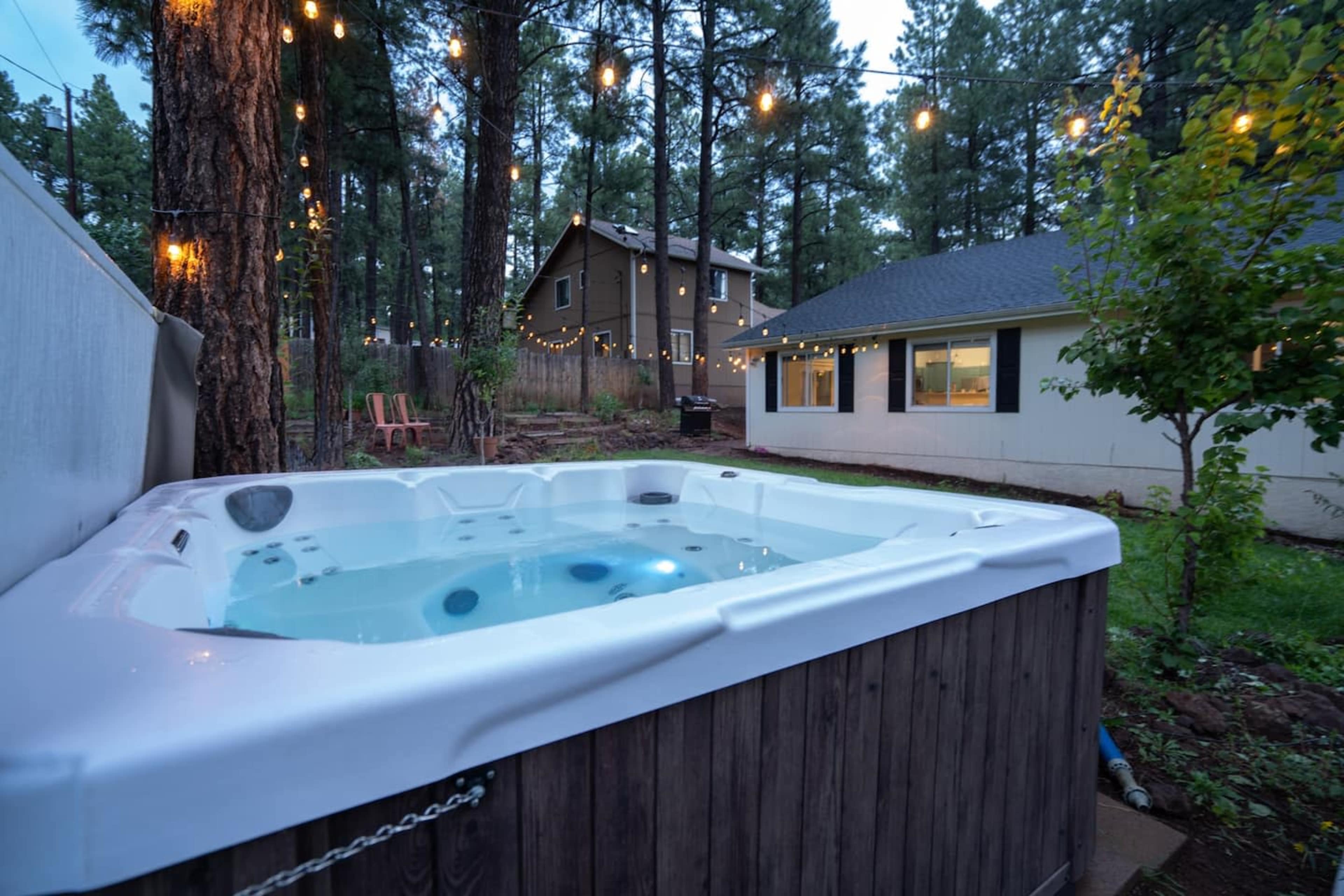 A hot tub is set in a backyard surrounded by tall trees and string lights, with two houses visible in the background.